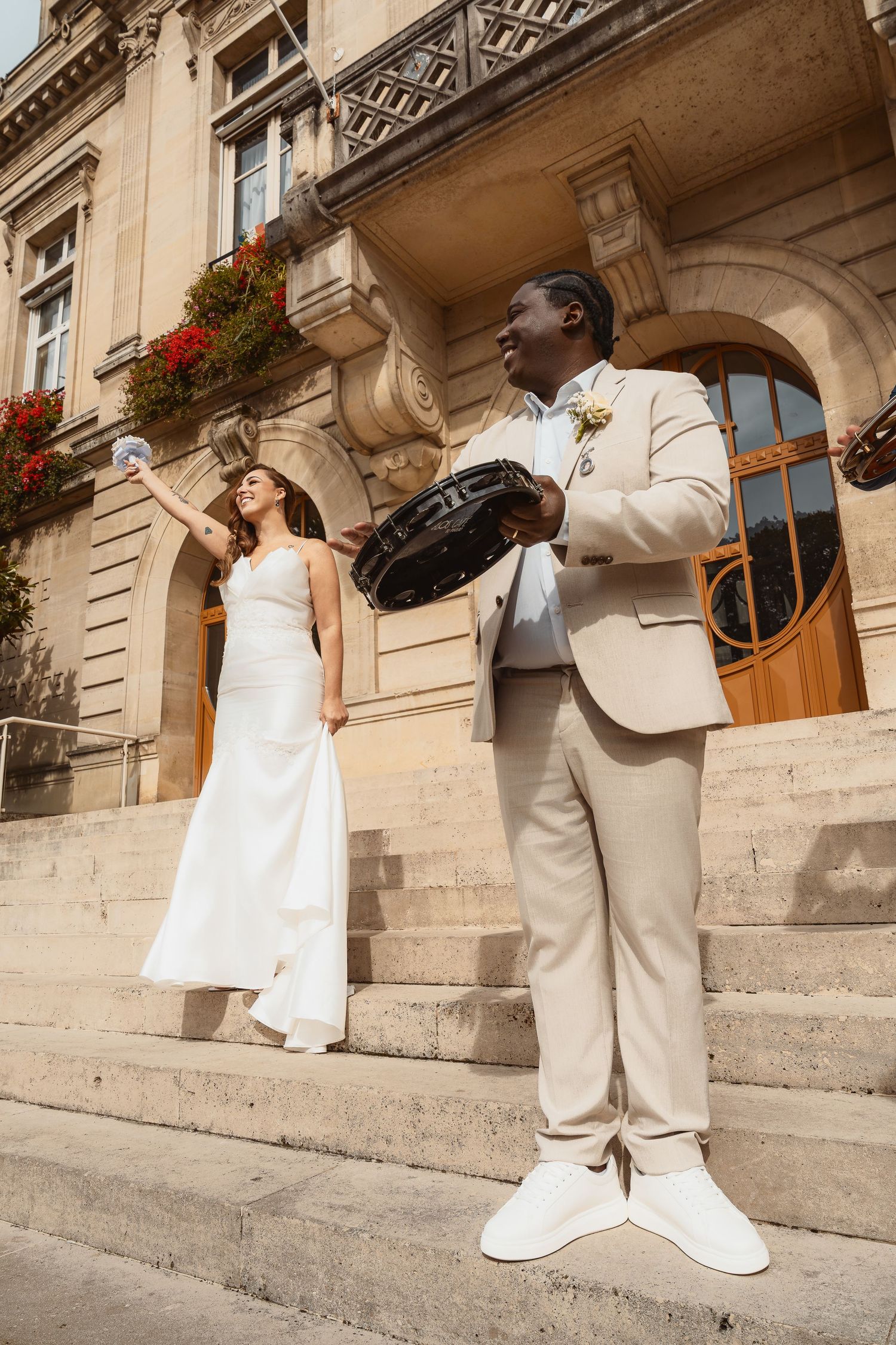 A bride and groom stand on stone steps outside an ornate European building with hanging flower baskets and arched doorways.