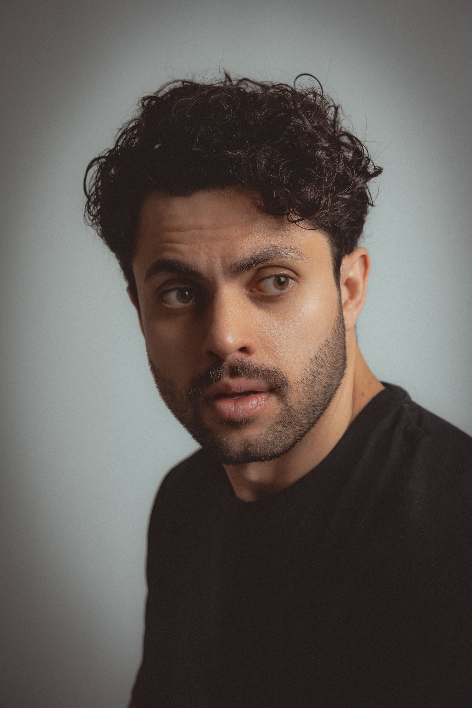 Studio portrait of a man with curly dark hair wearing a black t-shirt against grey background.