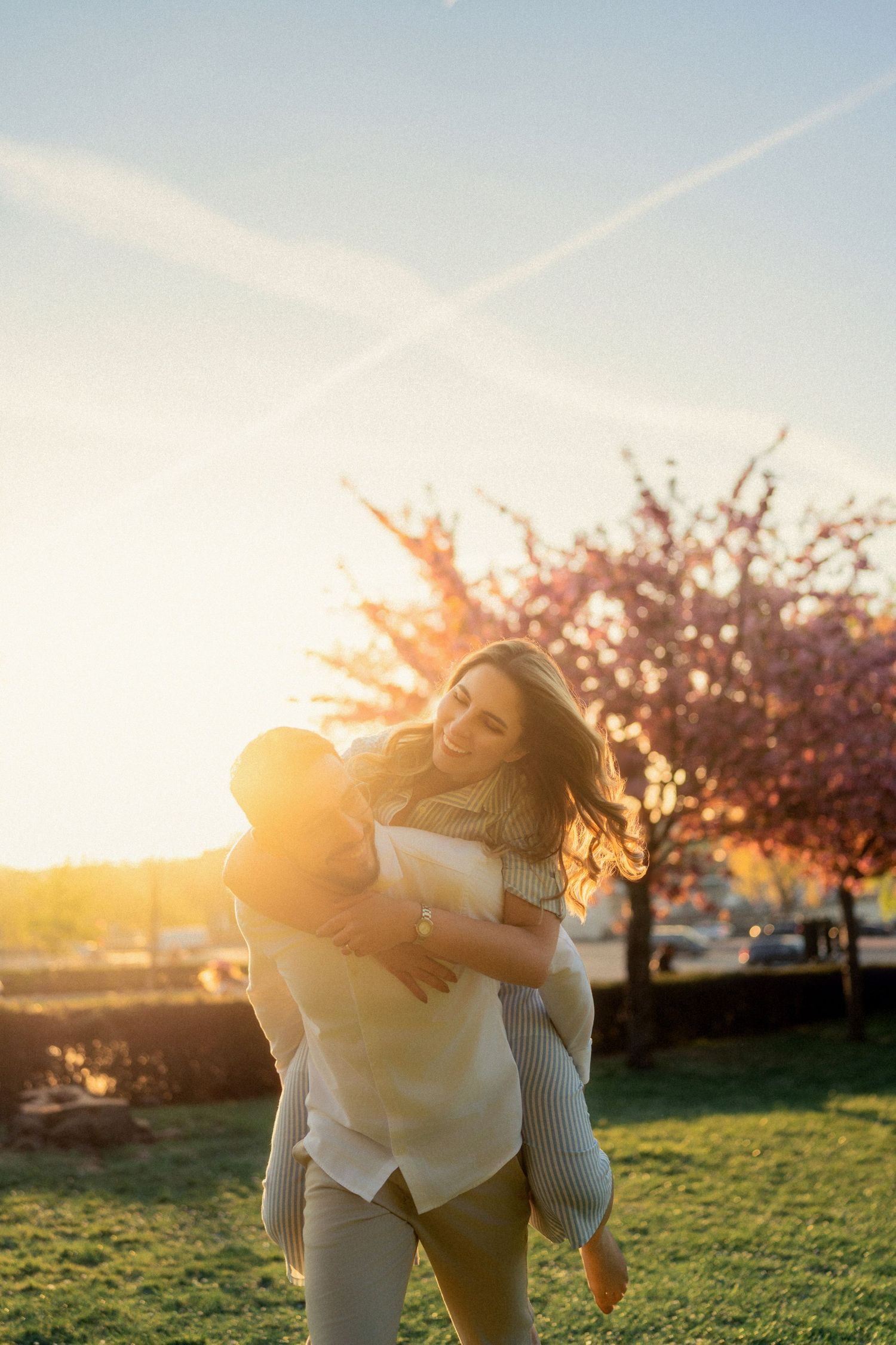 Two people embrace in a field at sunset with cherry blossom trees in the background creating a romantic silhouette.