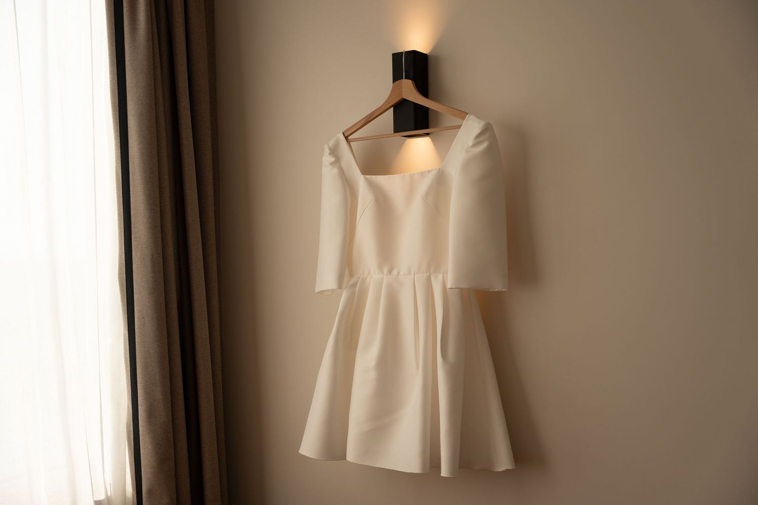 A white wedding dress hangs elegantly against a softly lit wall creating a romantic bridal preparation scene.