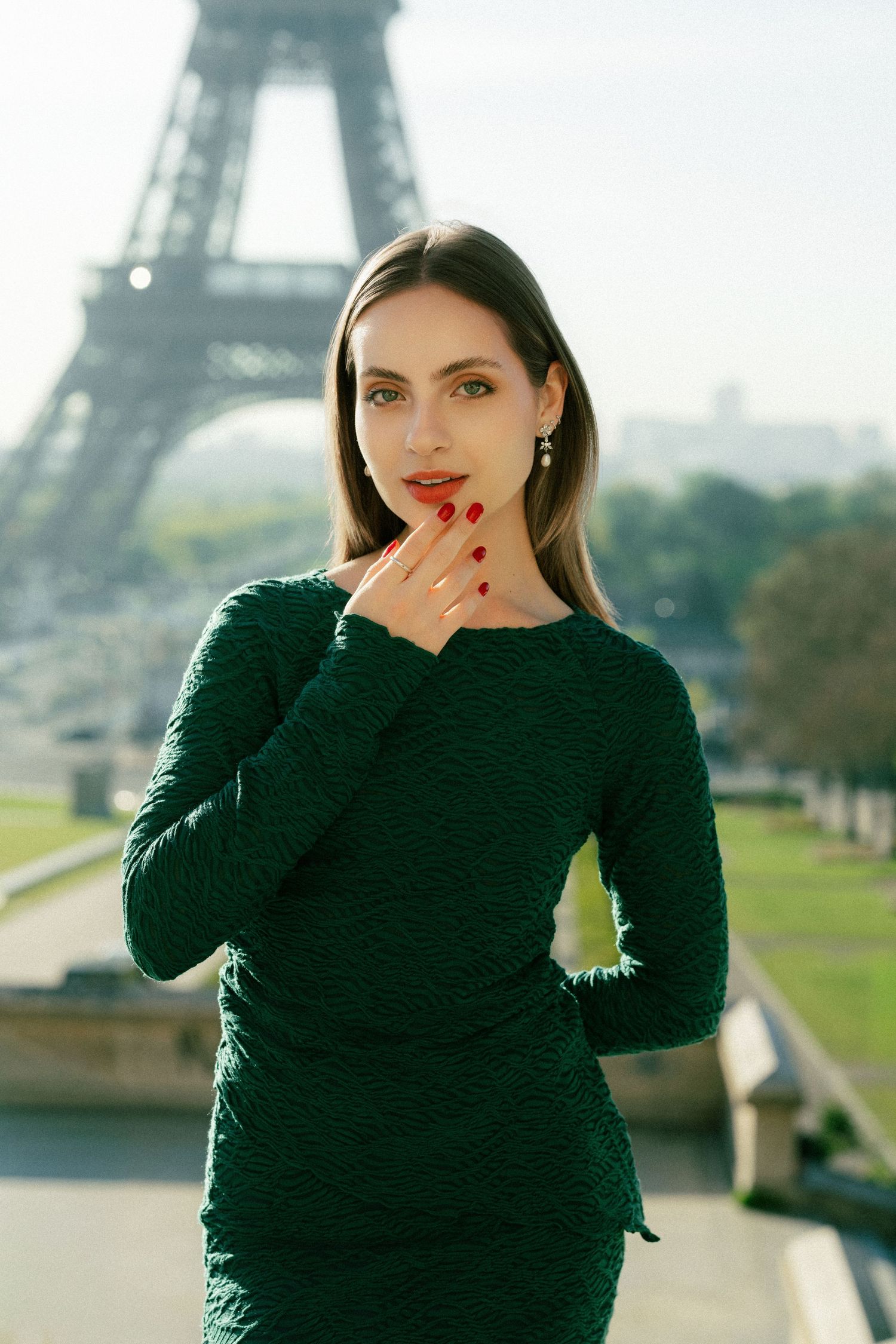 Elegant woman in green dress posing in front of the Eiffel Tower in Paris, with red nails and lips.