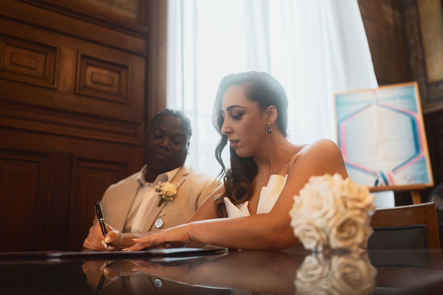 Bride signs marriage documents at ornate wooden desk with white roses in elegant indoor ceremony setting.
