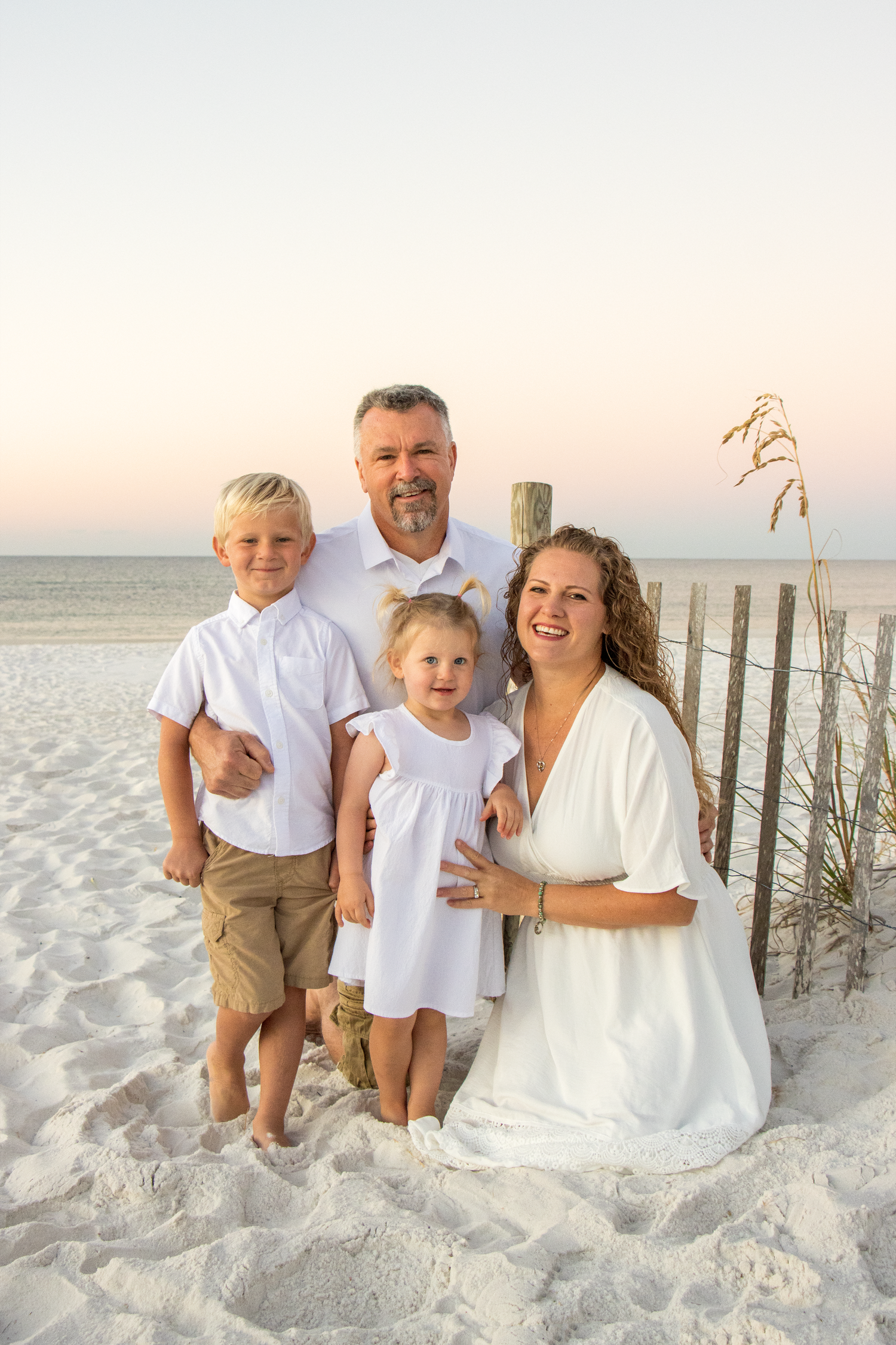 A family wearing white and khaki sits together on a sandy beach at sunset with wooden fence posts in the background.