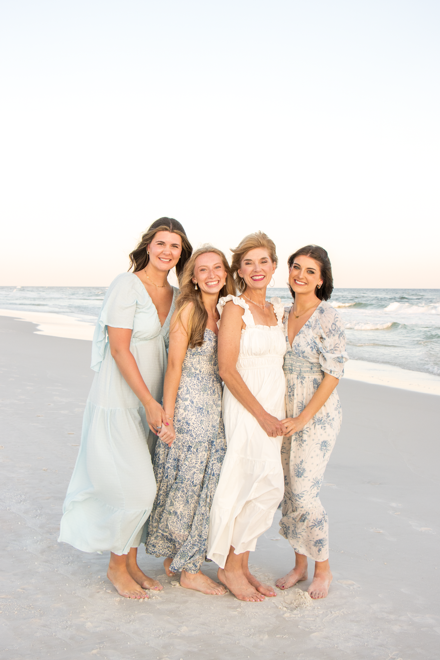 Group of friends in light summer dresses sitting together on white sand beach at sunset.