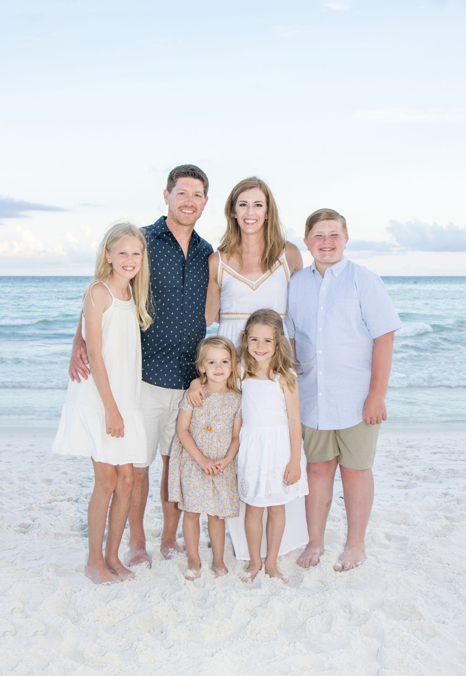 Beach family portrait with coordinated white and navy outfits against ocean backdrop.