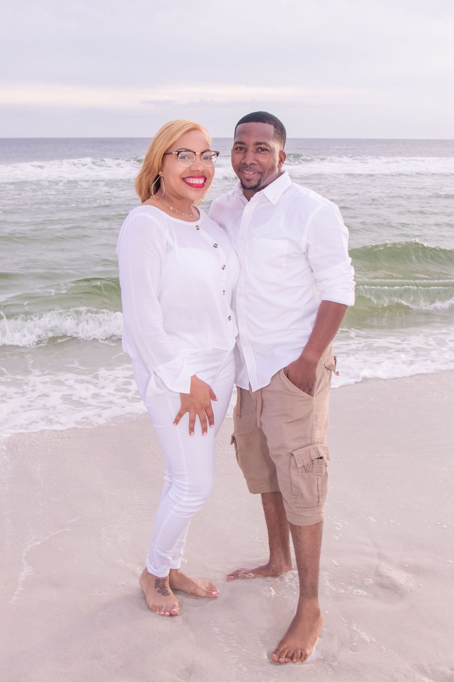 Couple in white outfits smiling together on beach with waves crashing behind them.