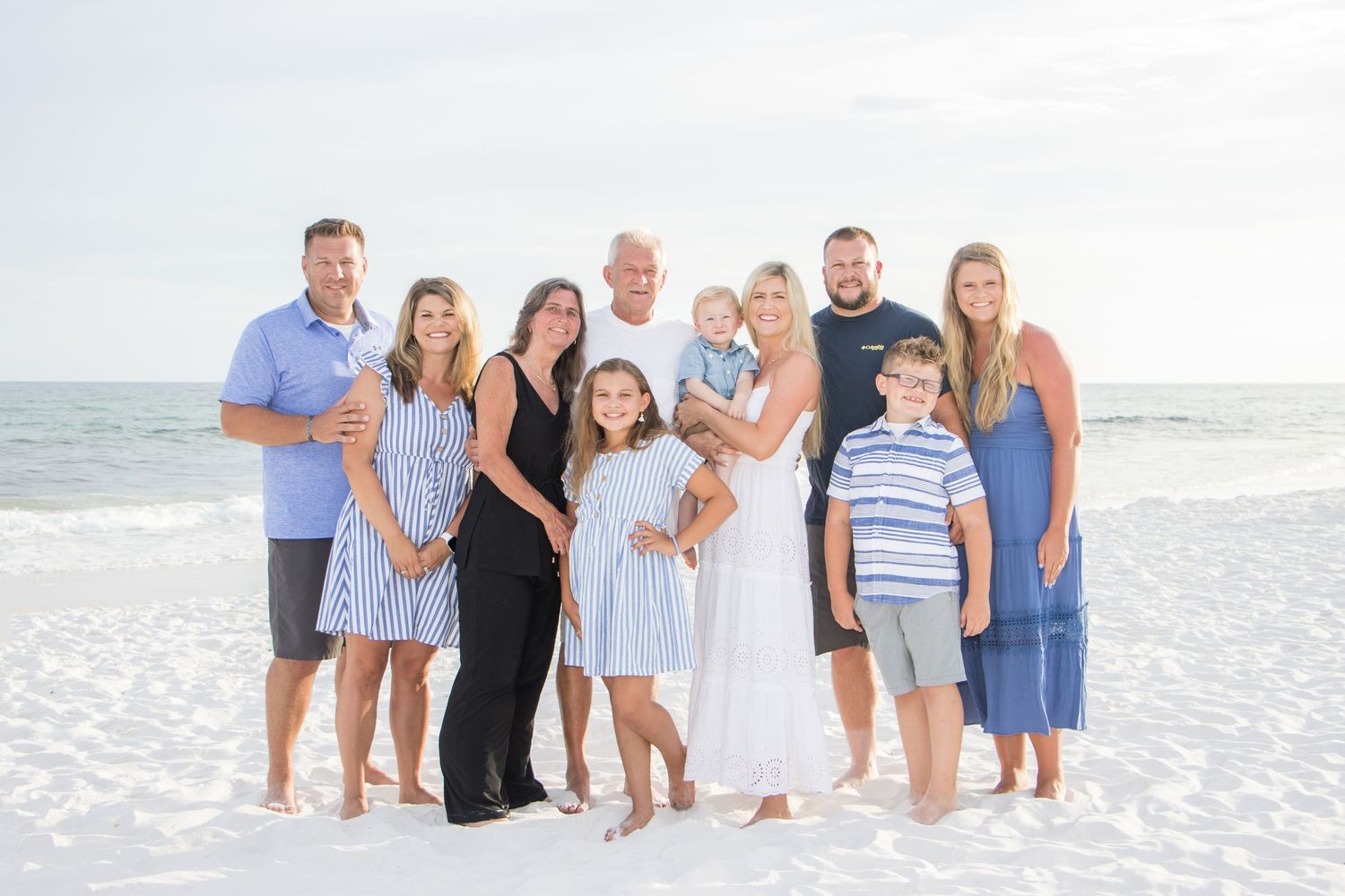 Extended family group wearing coordinated blue and white outfits poses together on a beach at dusk.