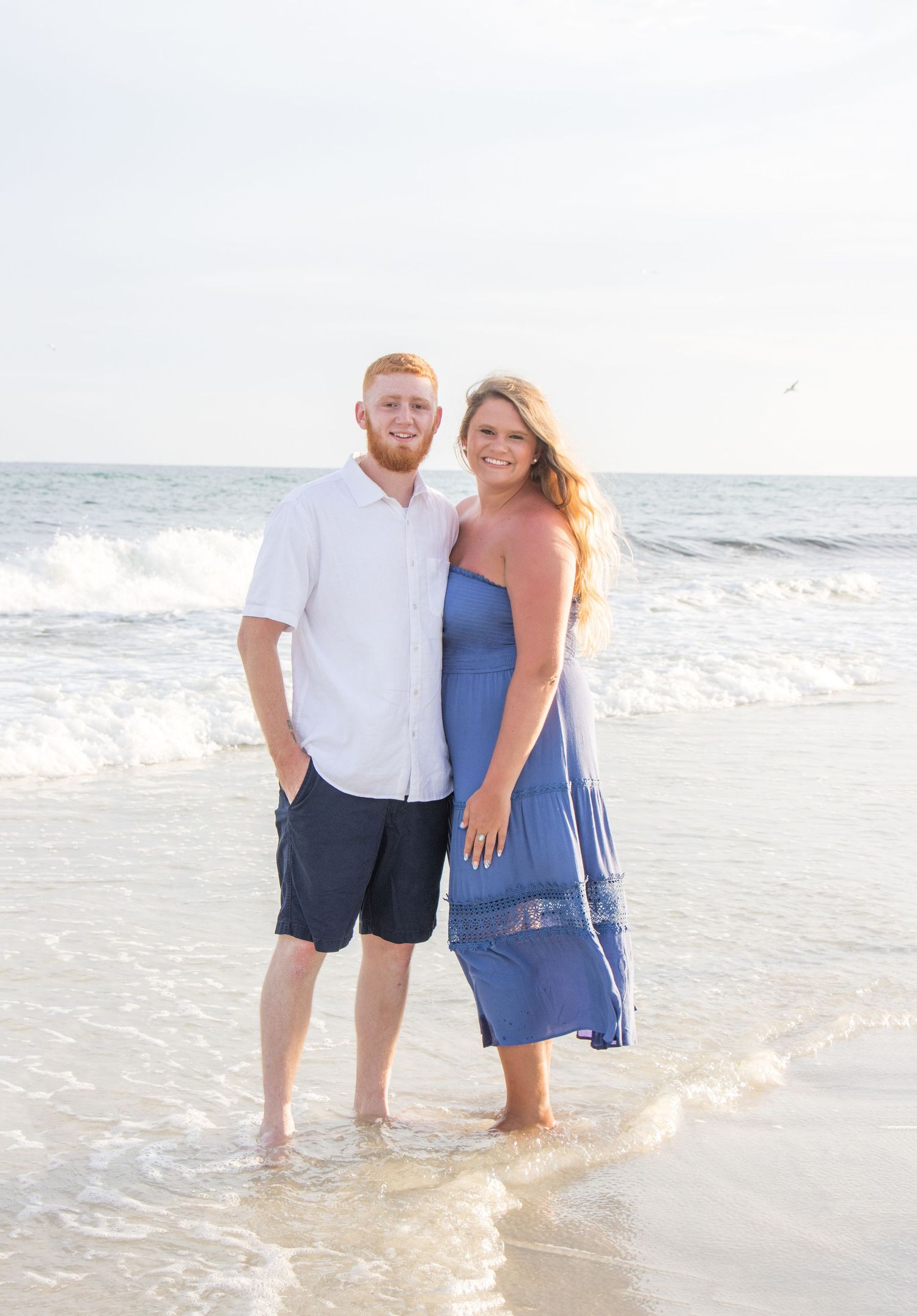 Couple in blue and white outfits standing together in ocean waves at sunset.