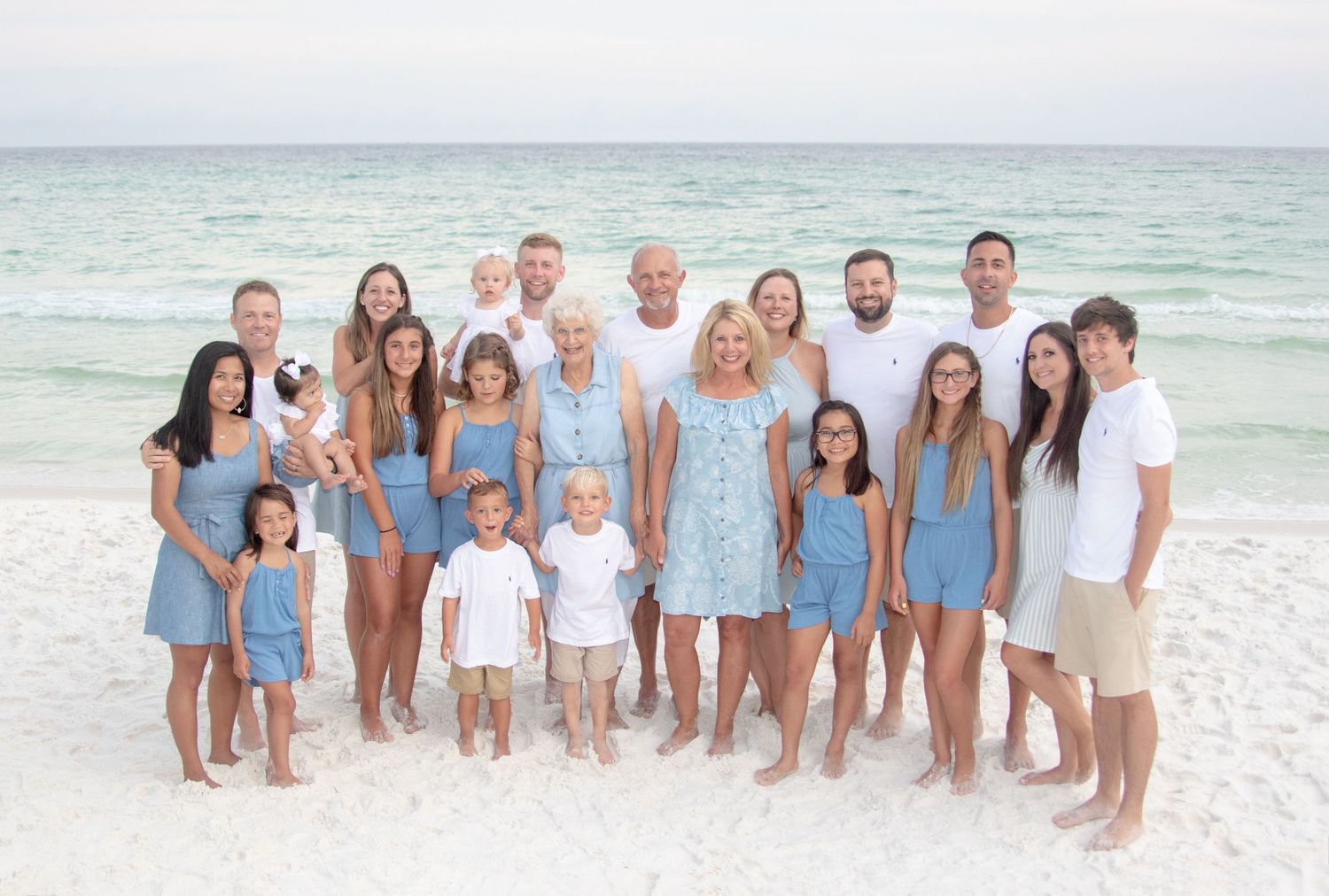 A group of people wearing coordinated white and blue outfits pose together on a sunny beach with ocean waves behind them.