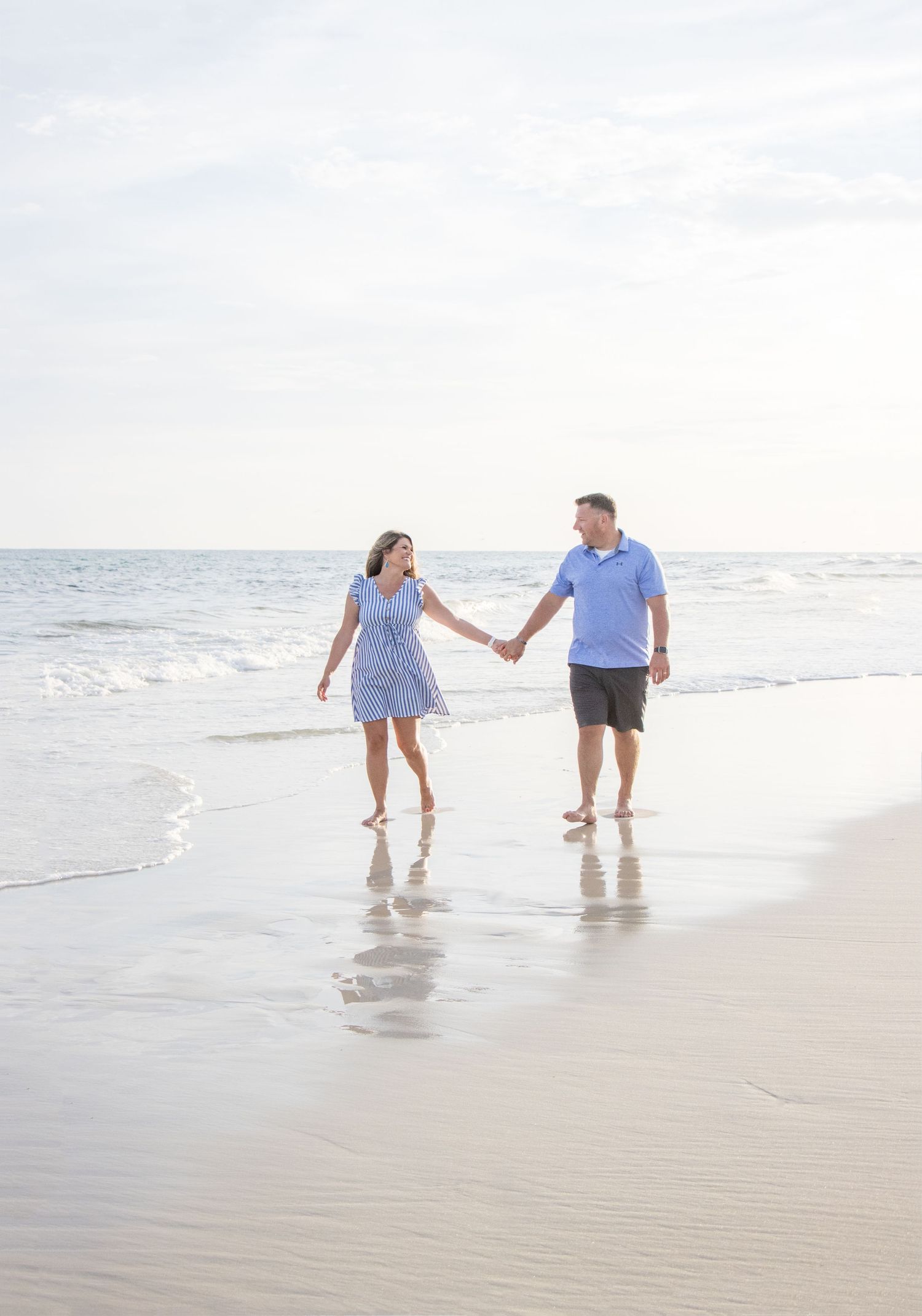 Couple holding hands while walking along shoreline of beach at sunset.