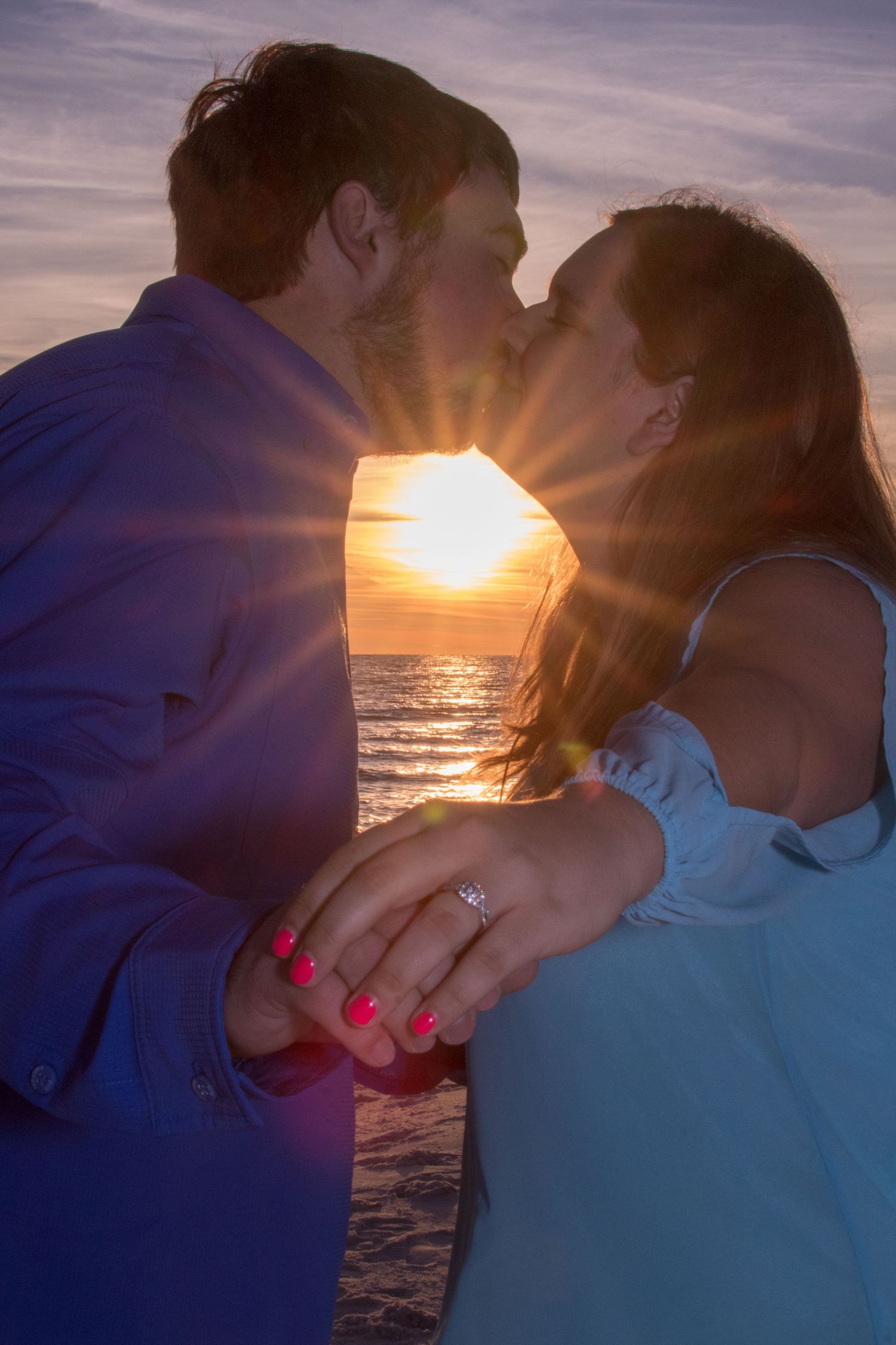 Romantic silhouette of couple kissing at sunset on beach with sun rays peeking through.