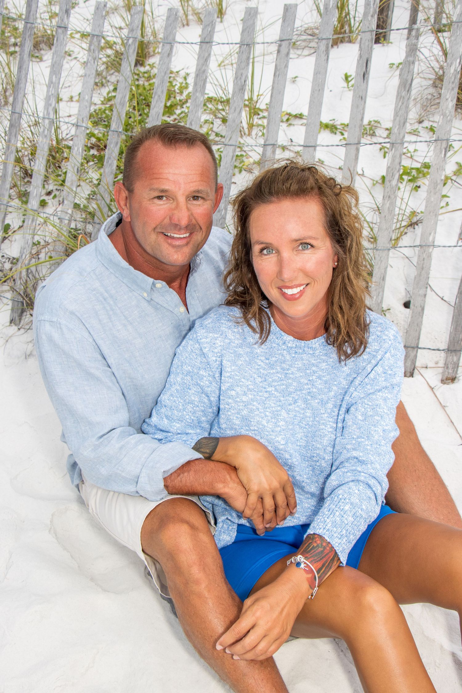Couple in casual blue attire sitting together by white beach fence.
