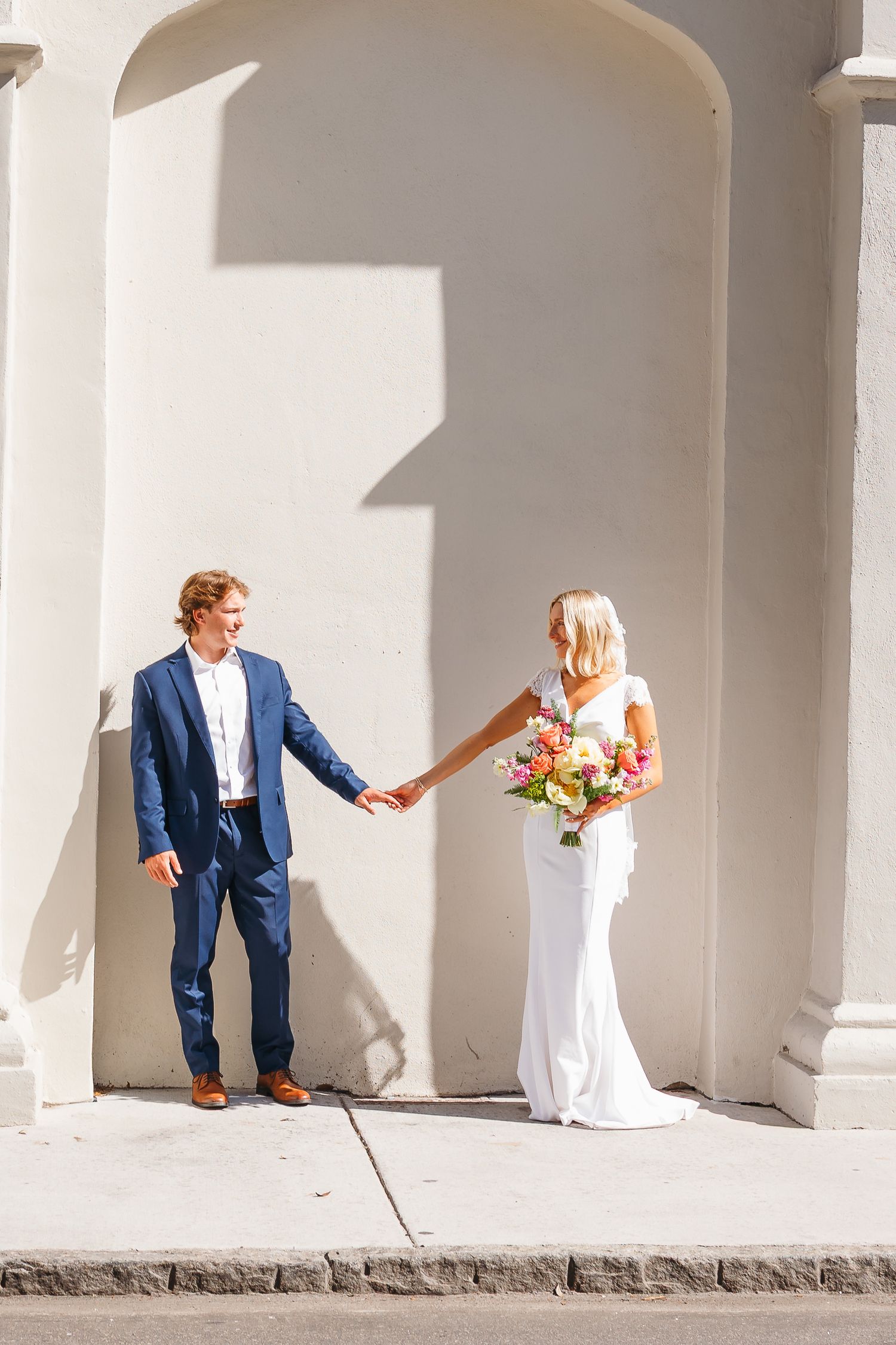 Bride in white gown holding colorful bouquet and groom in navy suit hold hands beside white arched columns.