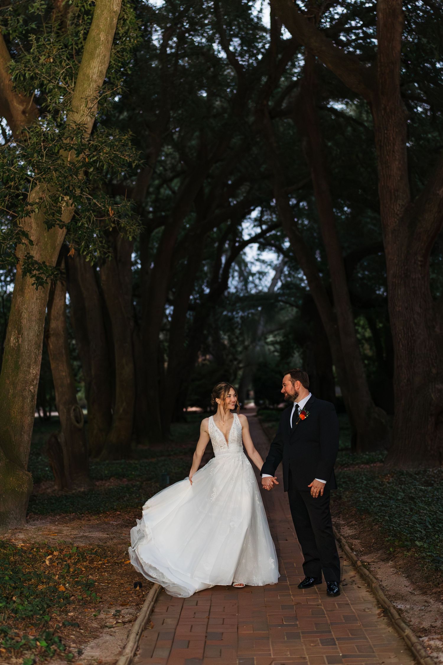Couple in wedding attire strolling along a romantic brick path surrounded by tall trees in a wooded setting.
