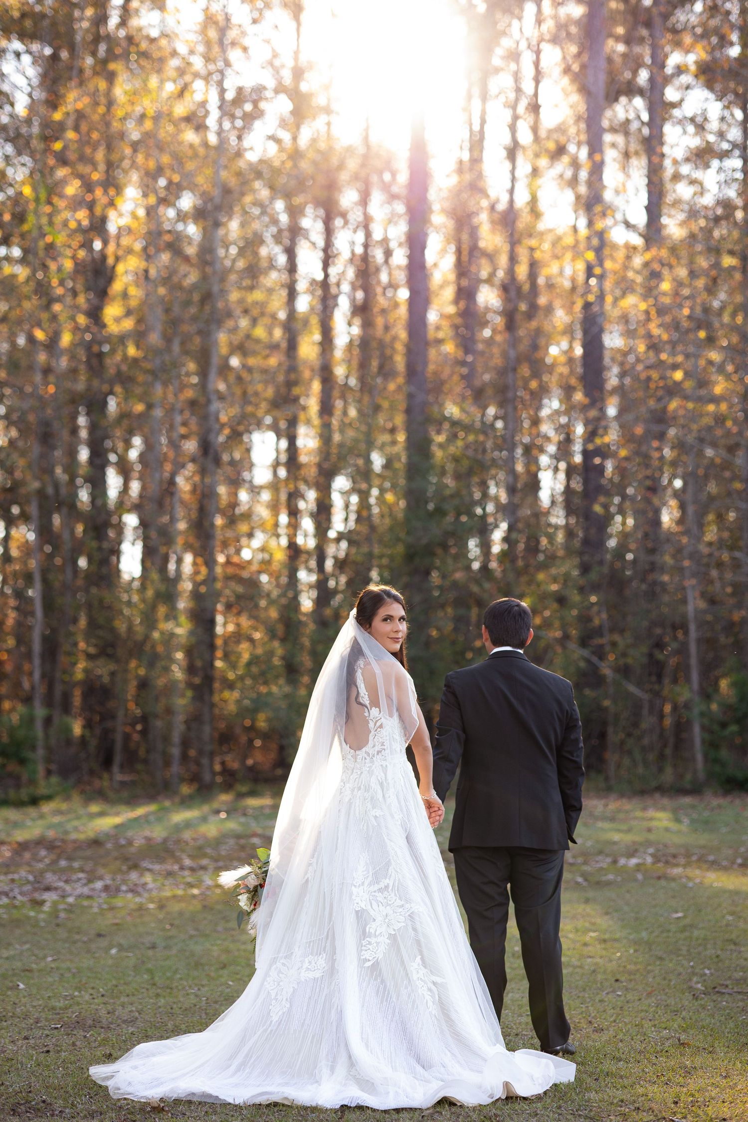 A couple in wedding attire walks through a sunlit autumn forest with golden leaves and tall trees.