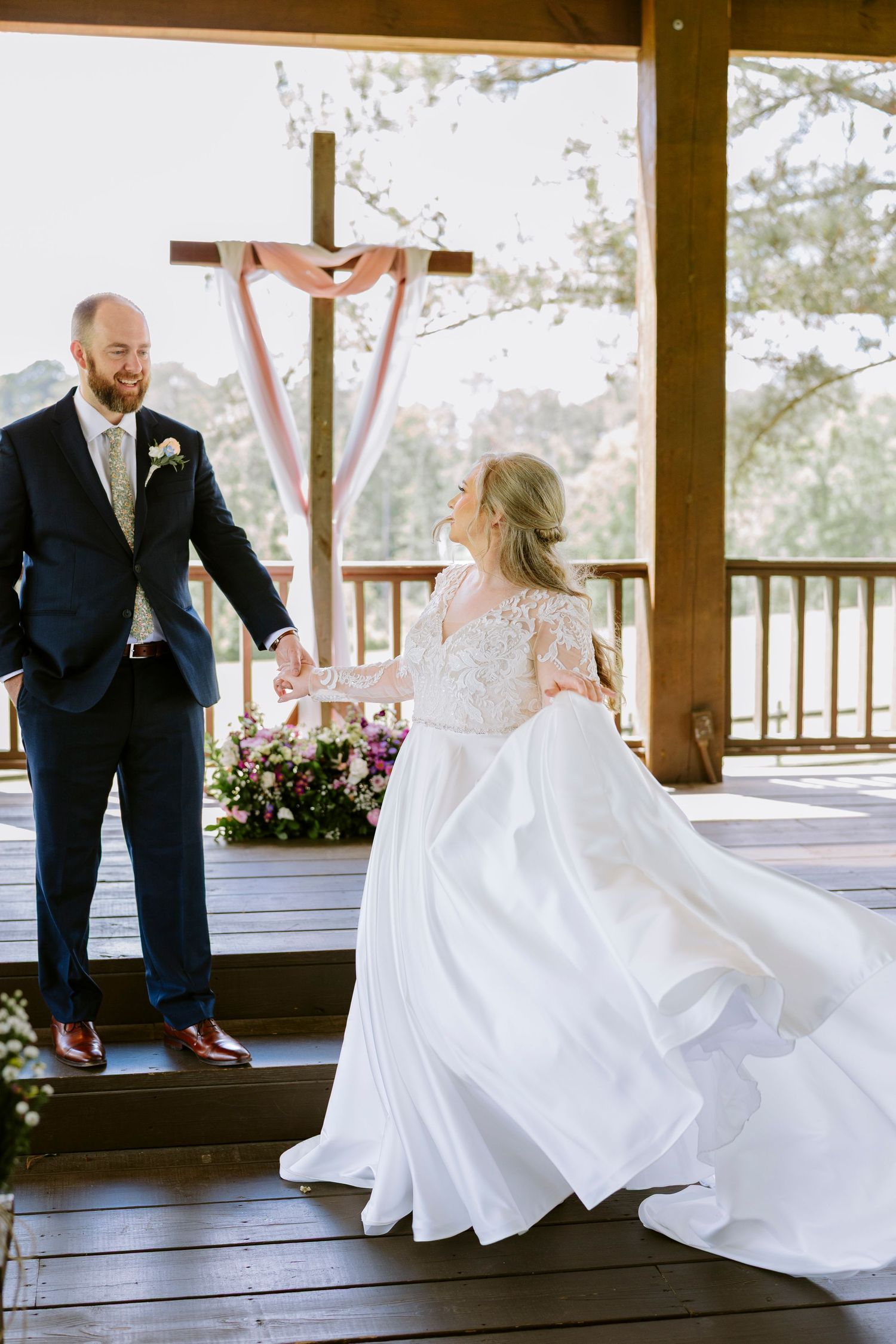 A couple in wedding attire share a playful moment on a wooden deck with scenic views in the background.