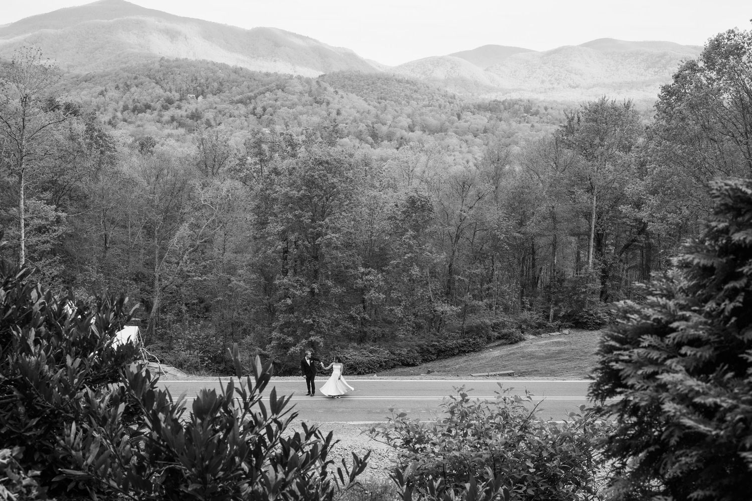 Black and white photo of a bride and groom walking on a path surrounded by lush forest and mountain views.
