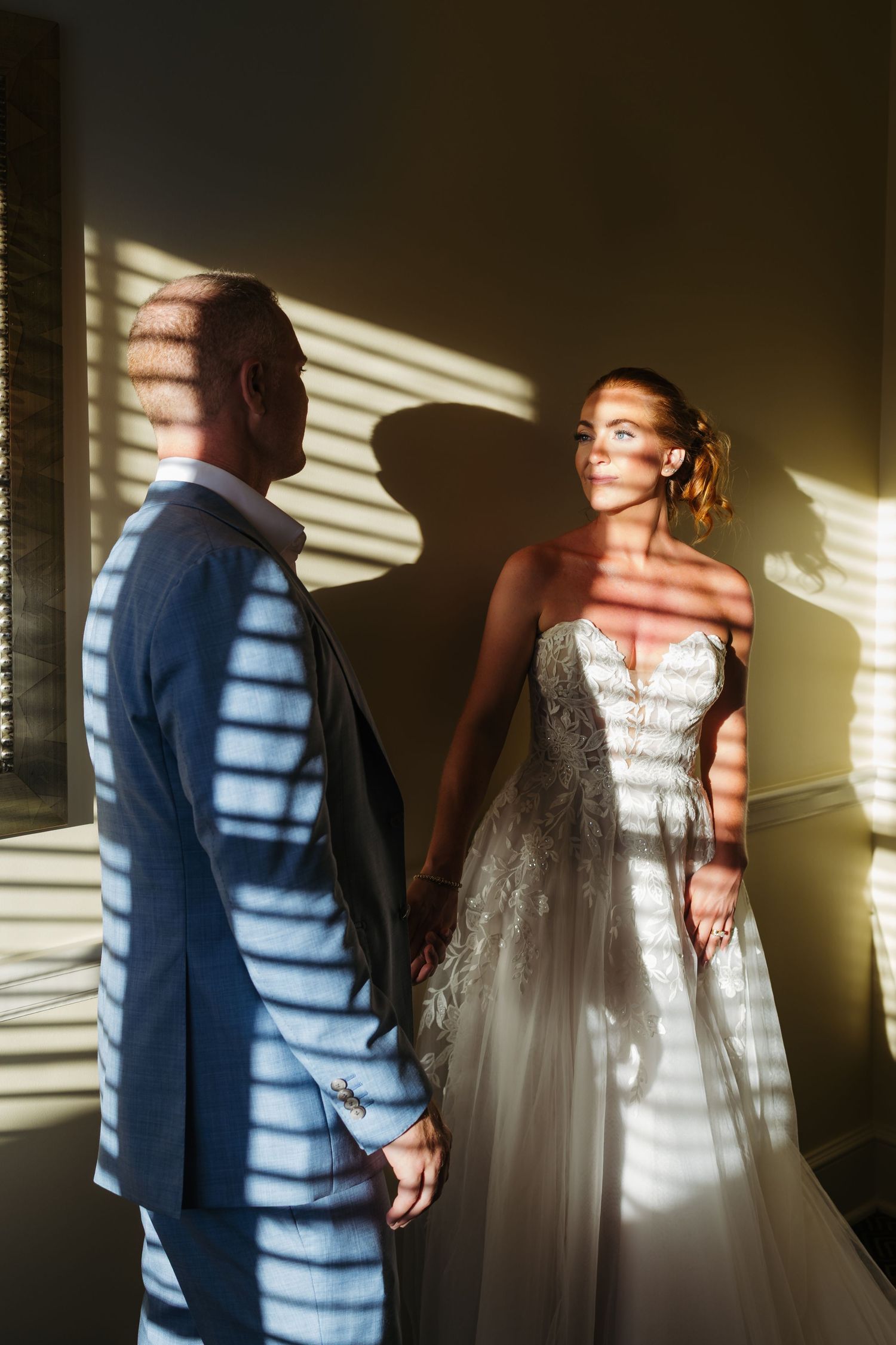 A bride and groom hold hands in dramatic striped light from blinds, casting bold shadows on a warm golden wall.
