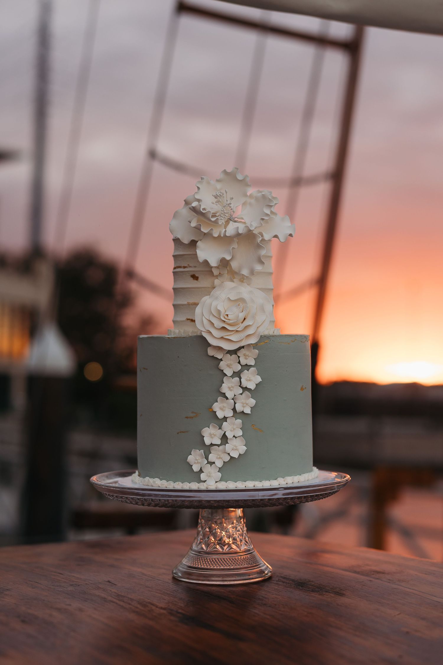 A sage green wedding cake with white sugar flowers against a sunset backdrop.