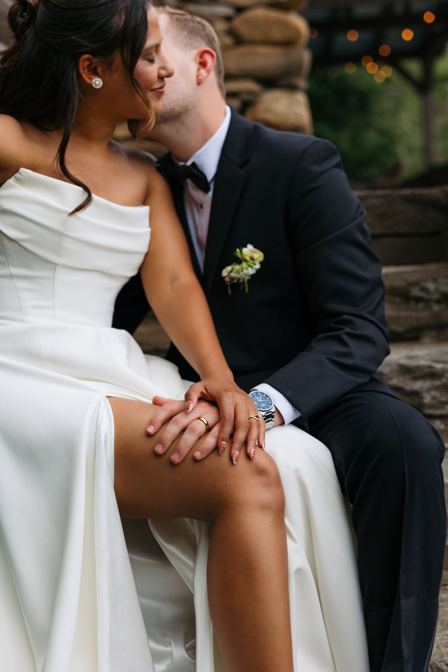 Bride and groom sitting close together holding hands, wearing wedding rings, in elegant wedding attire outdoors.