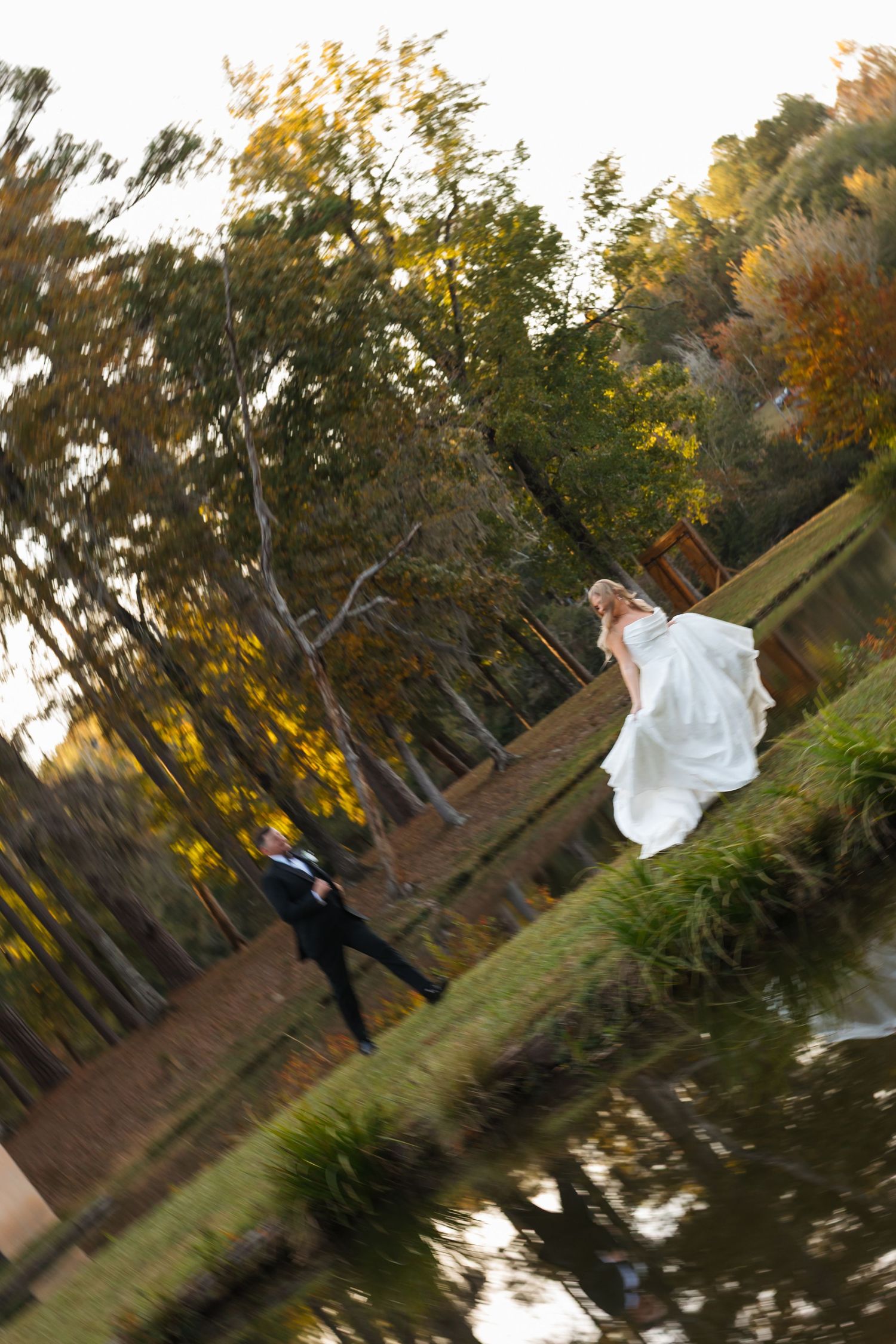 Bride and groom walking by a reflective pond surrounded by autumn trees in a romantic outdoor wedding photo.