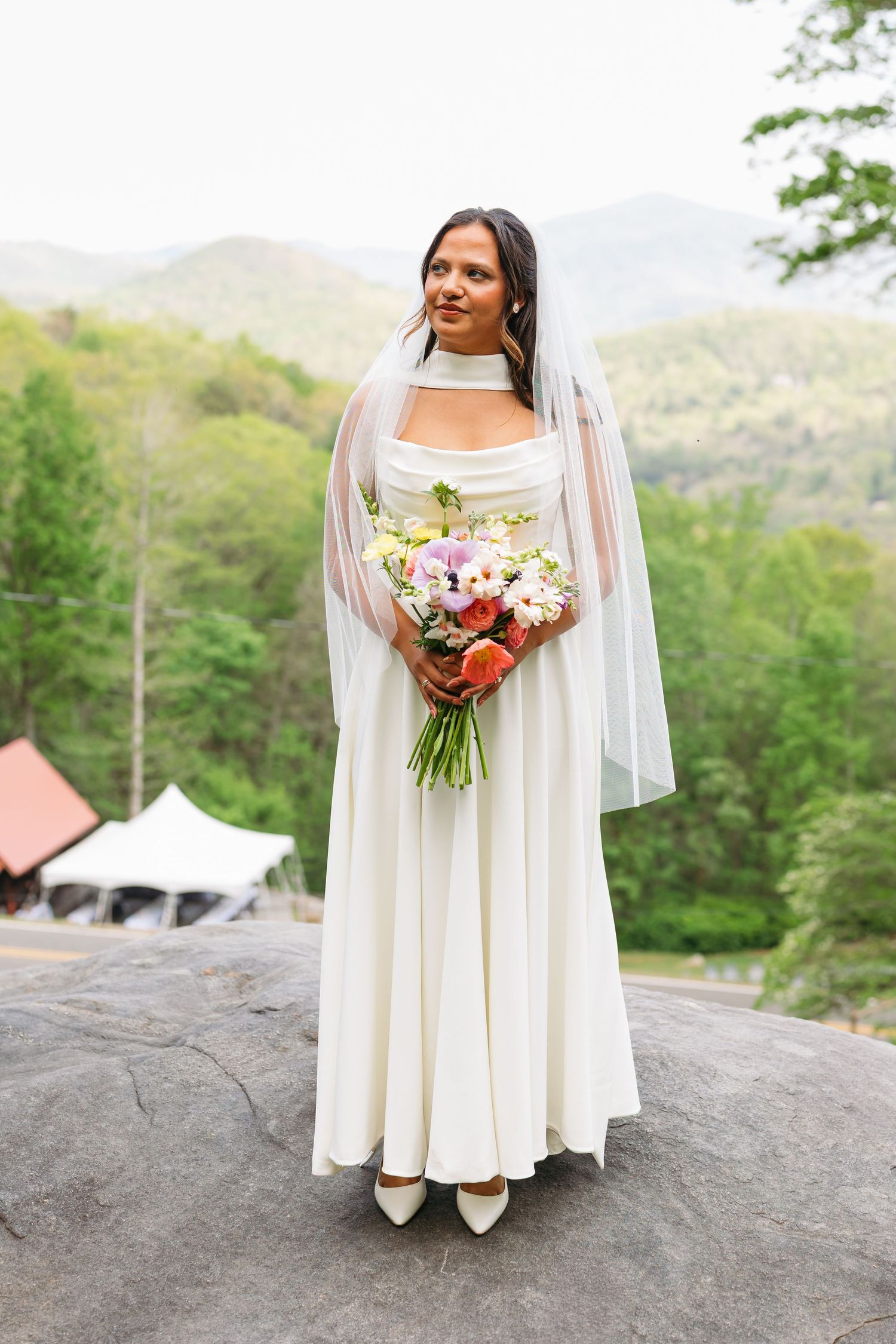 Bride in white gown and veil holds colorful bouquet on rock with mountain backdrop.