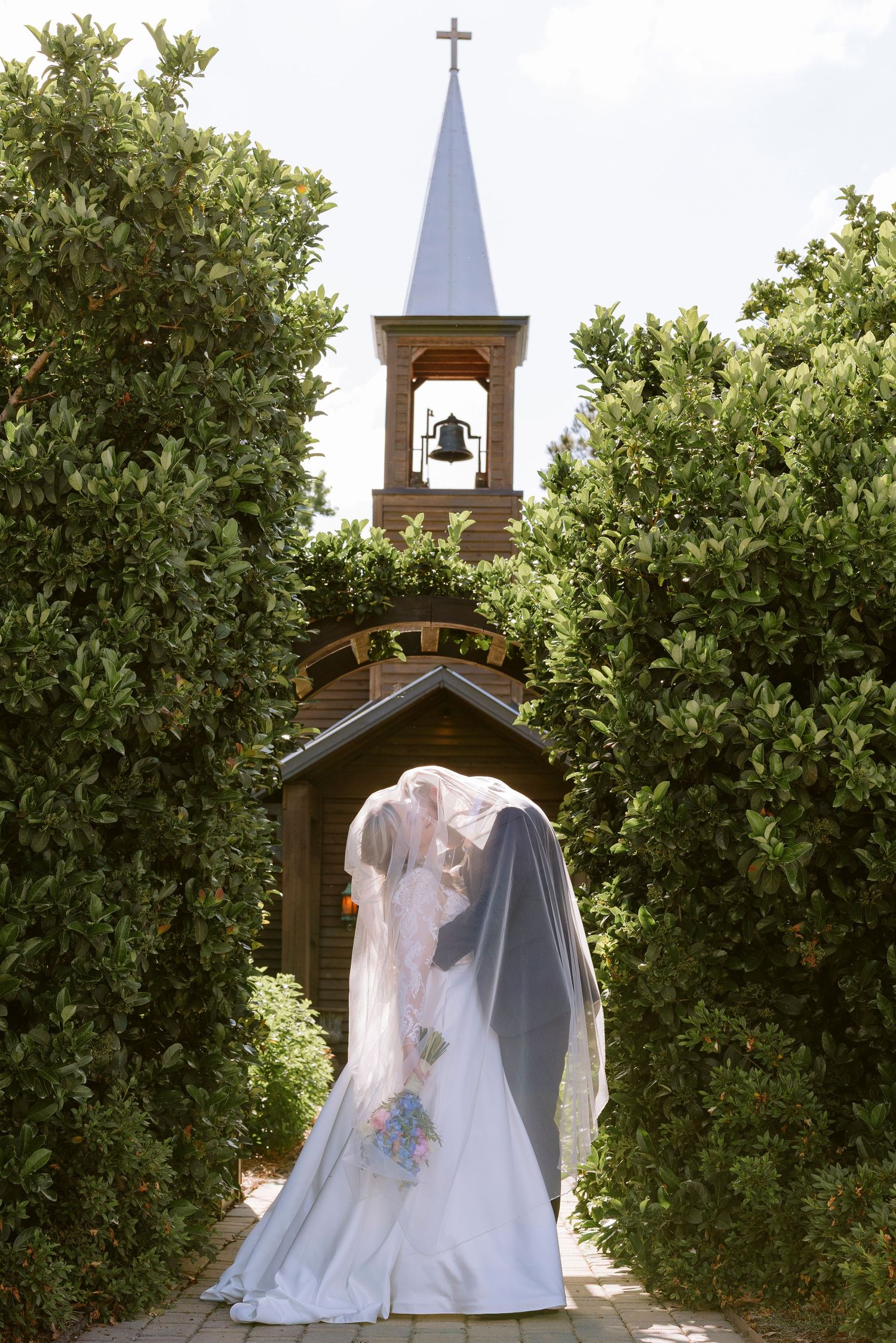 A wedding couple stands in front of a small chapel with a bell tower and steeple surrounded by lush green trees.