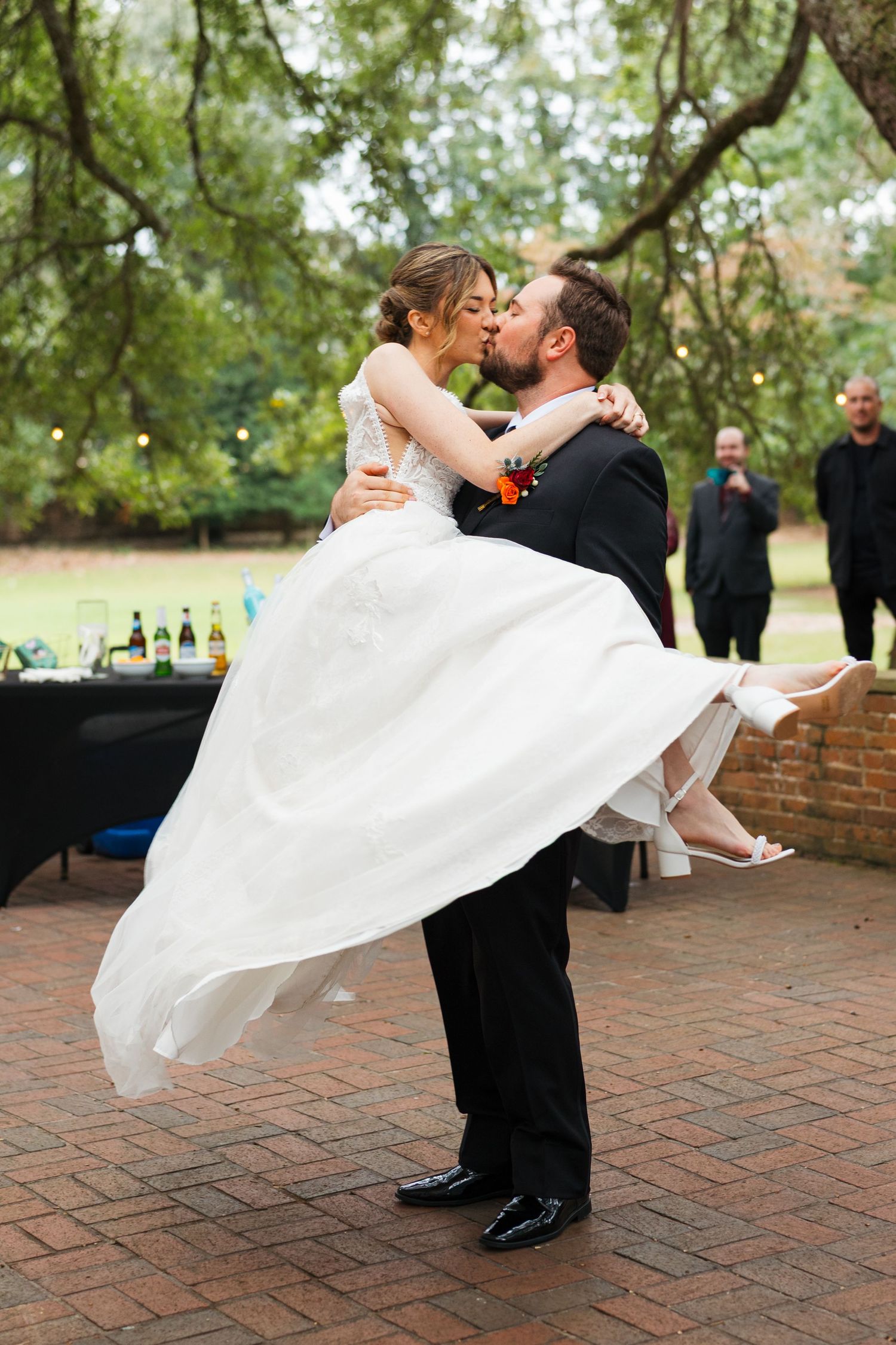 A romantic wedding couple shares a kiss as the groom dips the bride in her flowing white dress on a brick patio.