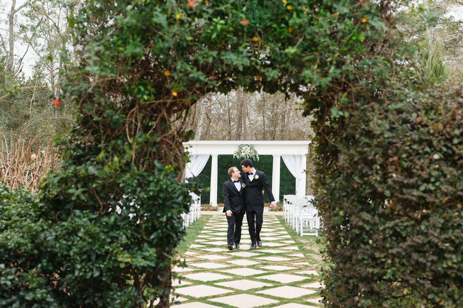 Two people walk along a garden path with a geometric paving pattern surrounded by lush greenery and hedges.