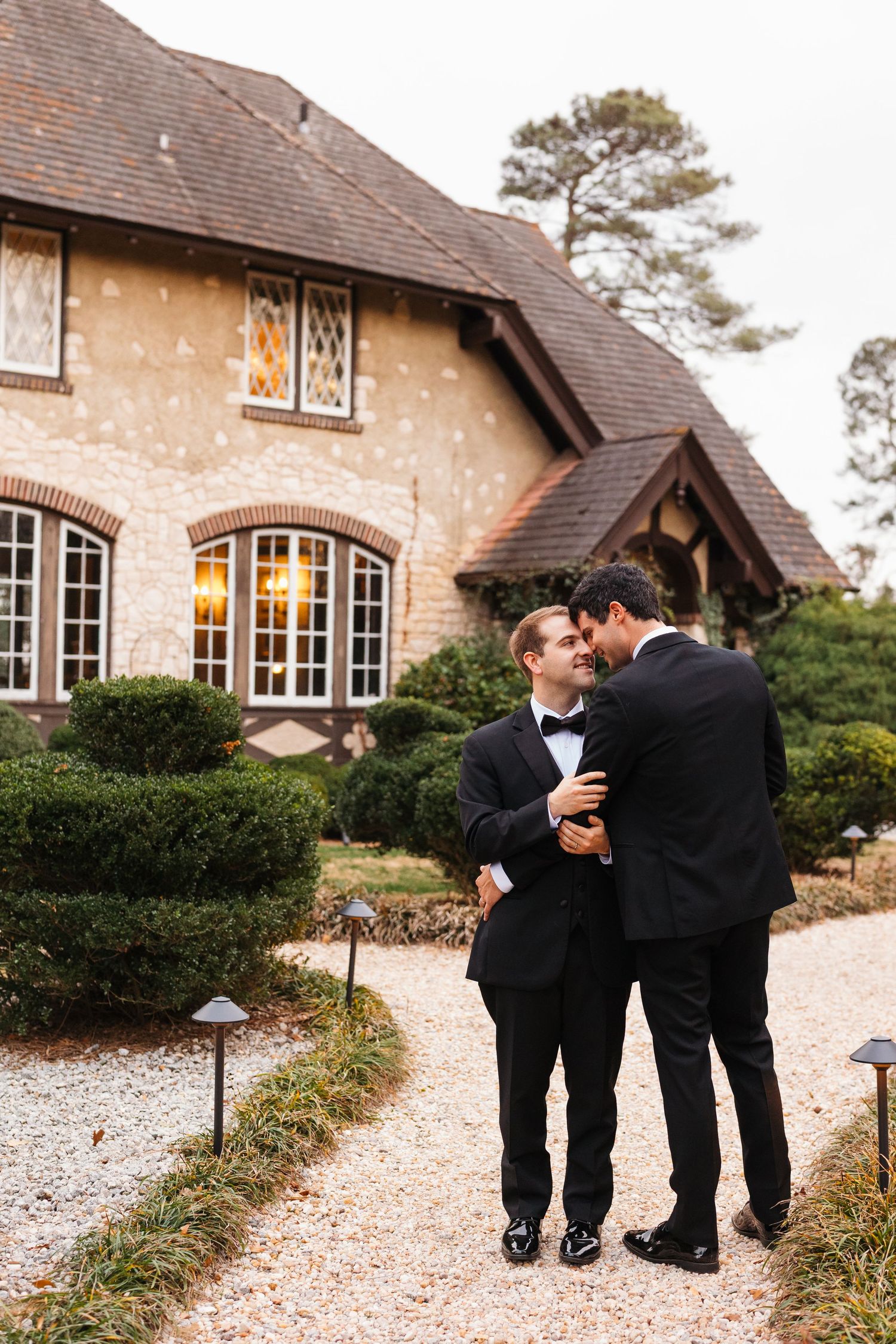 A romantic couple in formal black attire share a tender moment on a gravel path outside a rustic stone building.