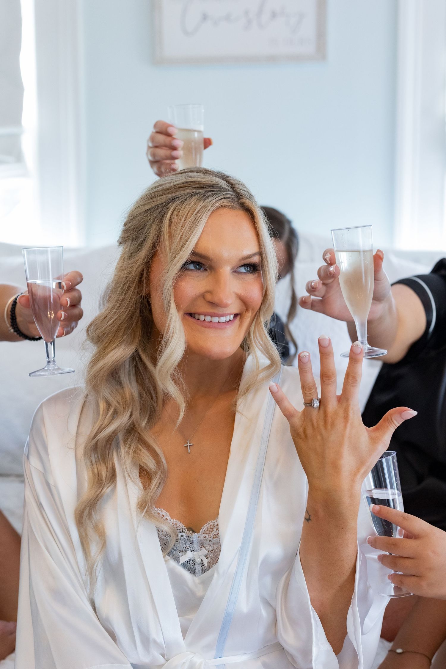 Bride in white robe holds up champagne glass during pre-wedding celebration with friends.
