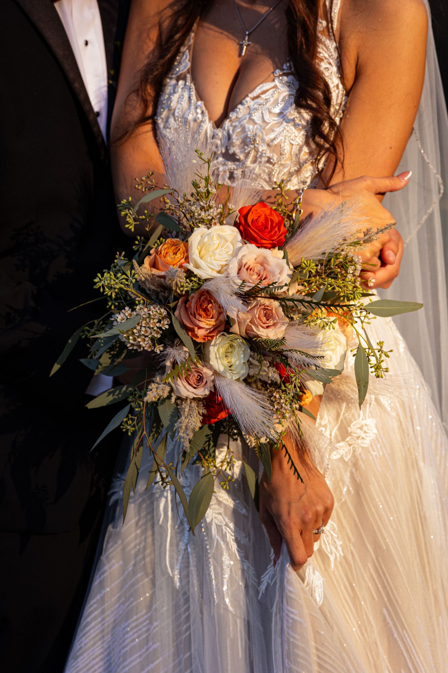 Wedding bouquet with blush roses, red flowers and greenery held against a white wedding dress in natural light.
