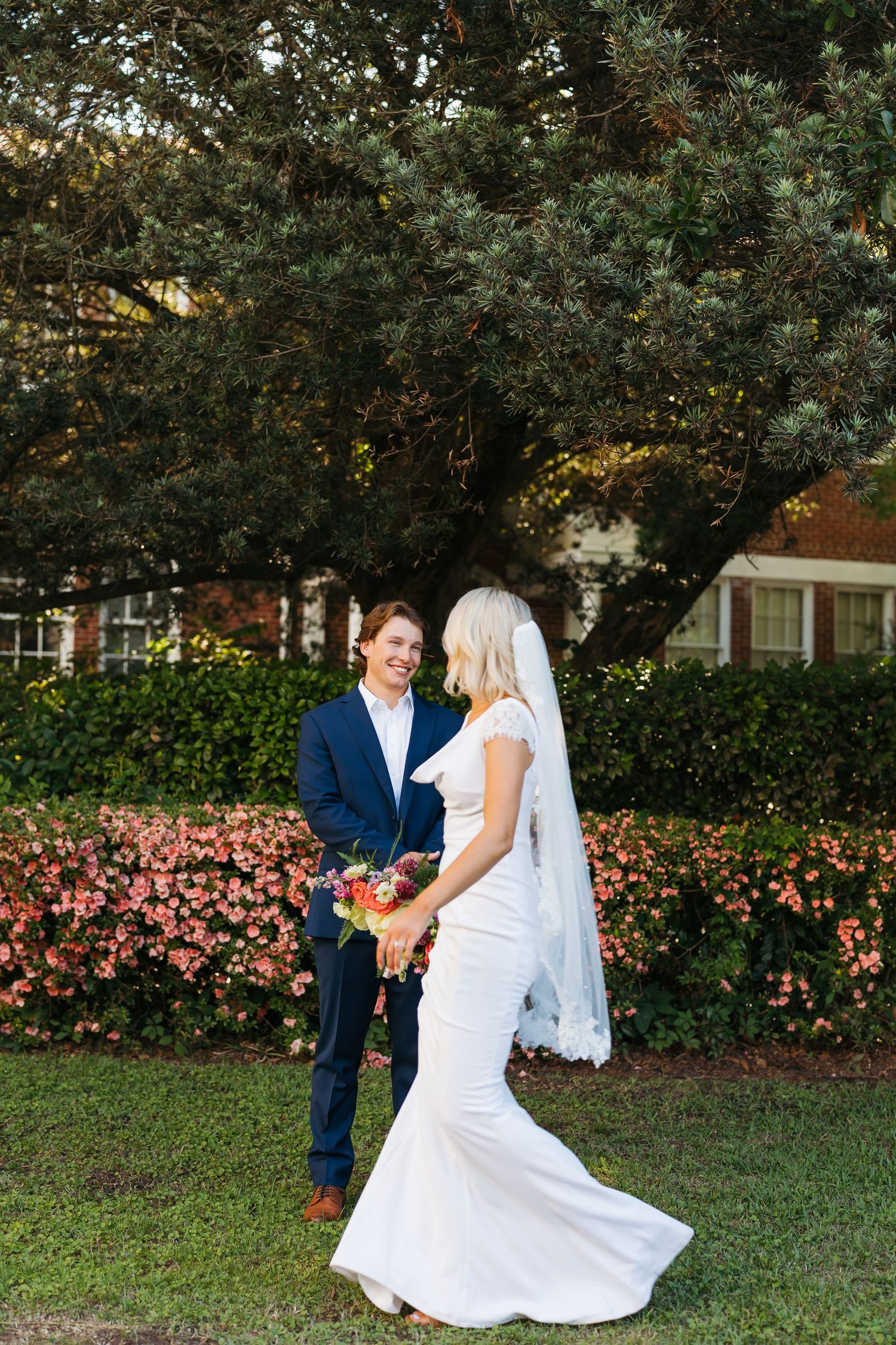 Bride in white gown holding colorful bouquet stands with groom in navy suit outdoors near lush greenery and pink flowers.