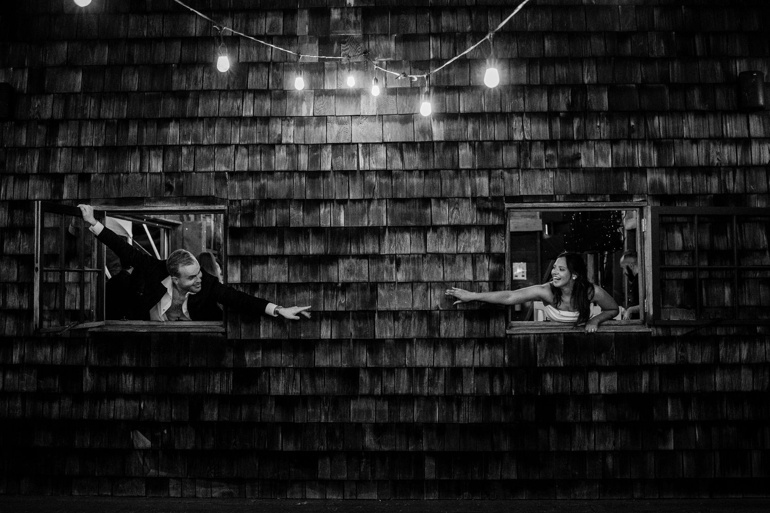 Black and white wedding photo of couple playfully posing in rustic barn windows under string lights at night.