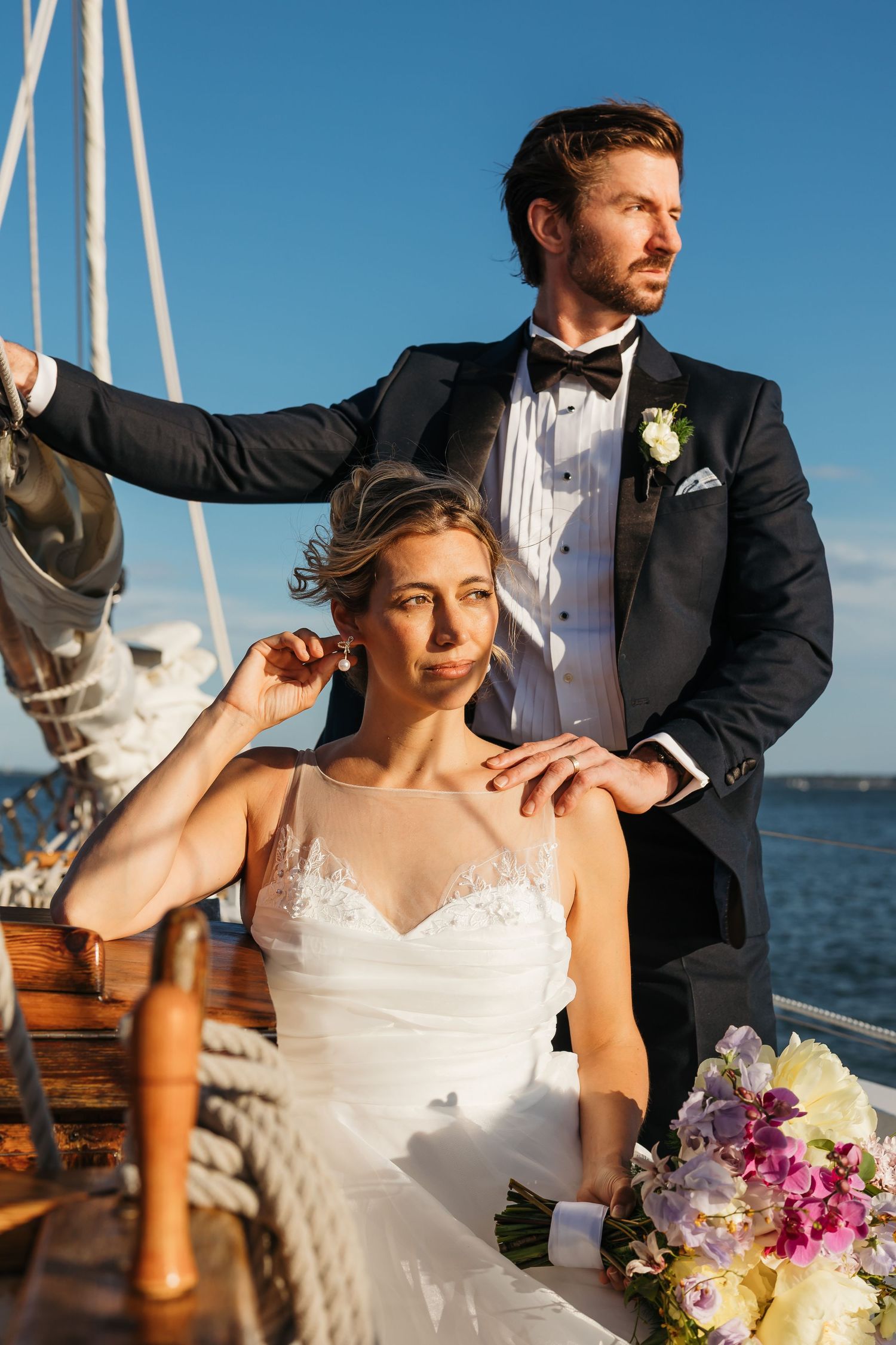 Bride and groom posing on a sailboat, bride in white gown holding bouquet, groom in black tuxedo, blue sky backdrop.