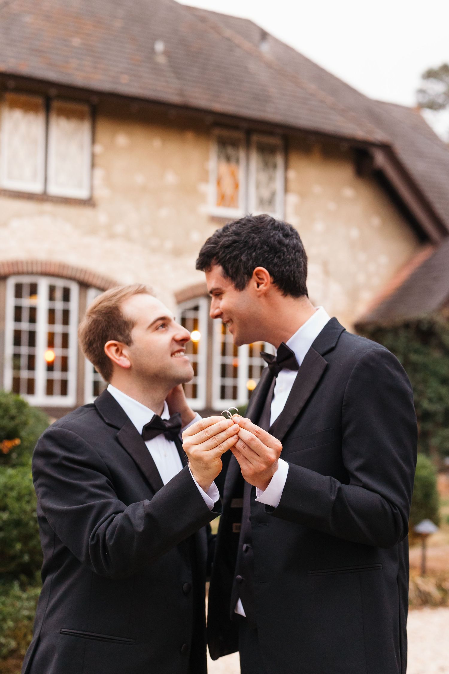 Two grooms in black tuxedos exchange rings outdoors in front of a elegant stone building on their wedding day.