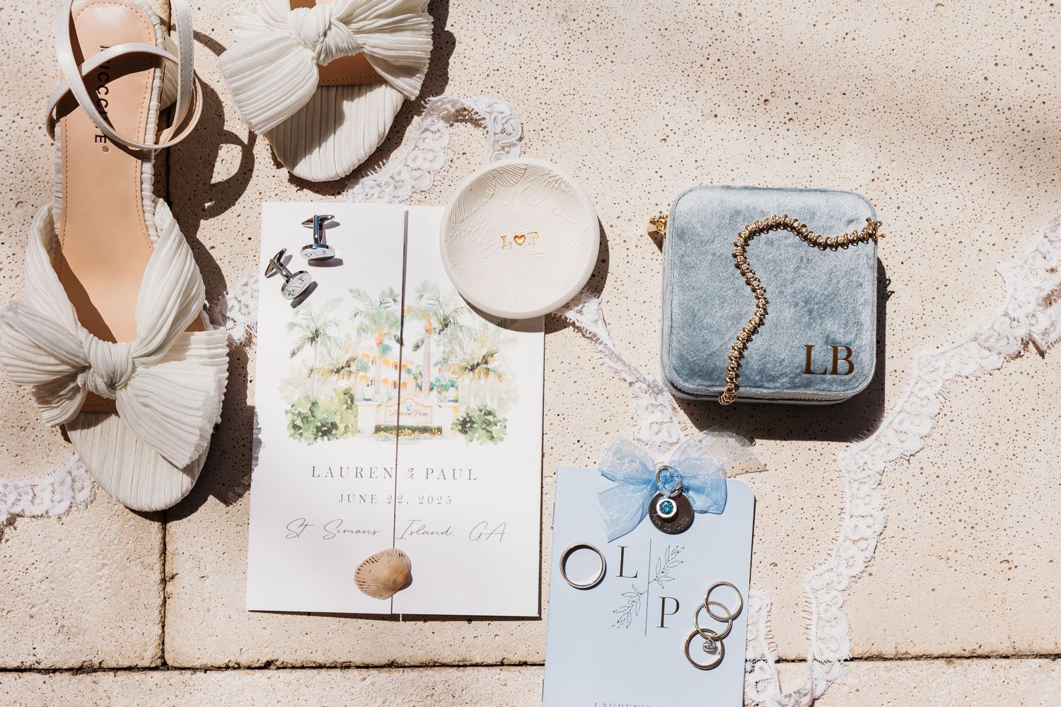 Flat lay of wedding invitation cards with sandals, candle, and blue ring box on light textured surface.