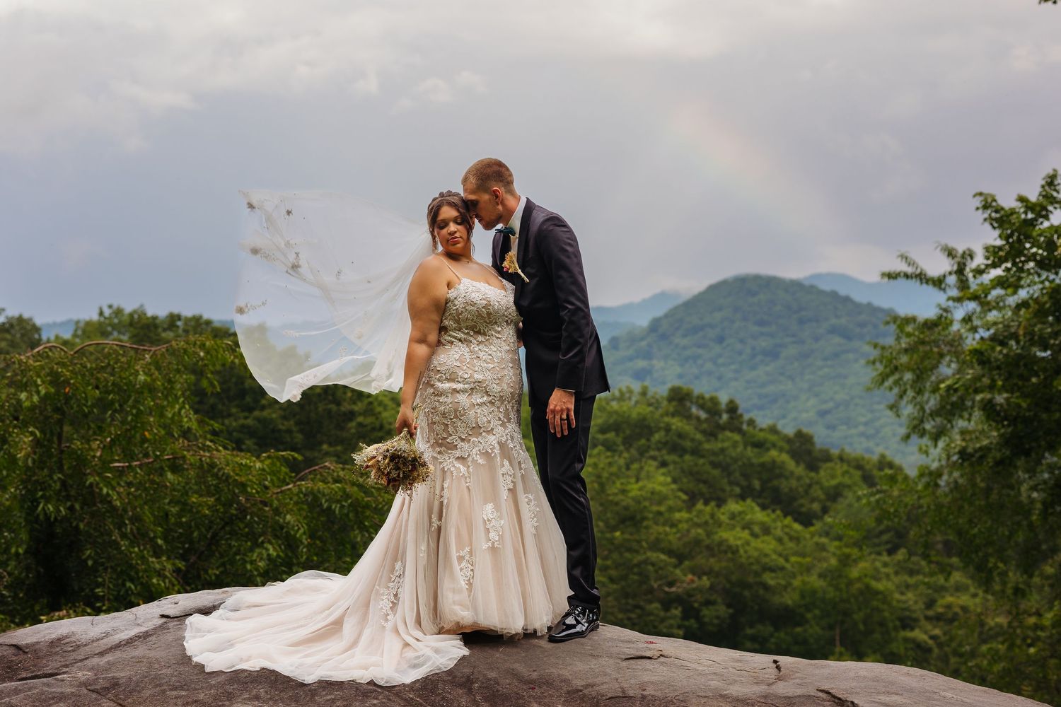 A couple in wedding attire shares an intimate moment on a scenic mountain overlook with lush greenery in the background.