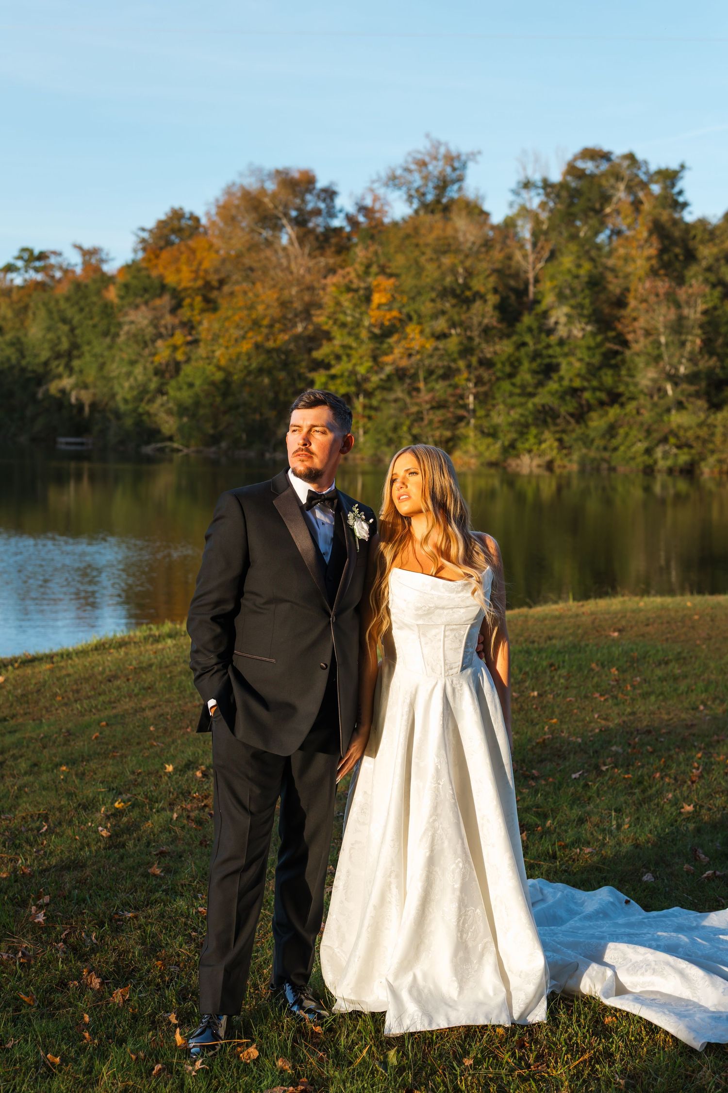 Bride and groom posing by a lakeside with autumn trees, bride in white gown, groom in black tuxedo.