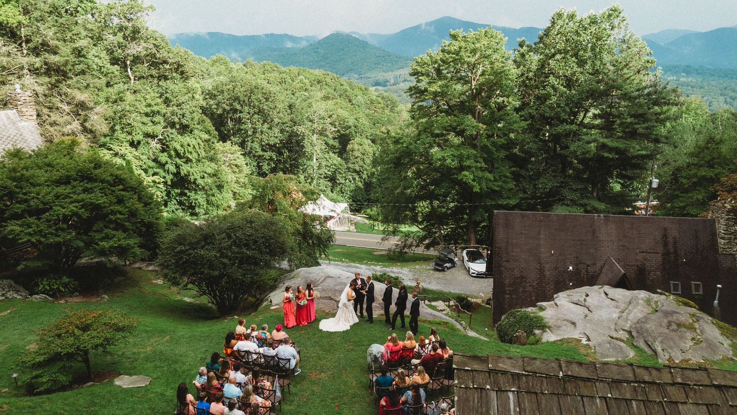 Panoramic view of a green mountain landscape with wedding ceremony setup and guests gathered outdoors.