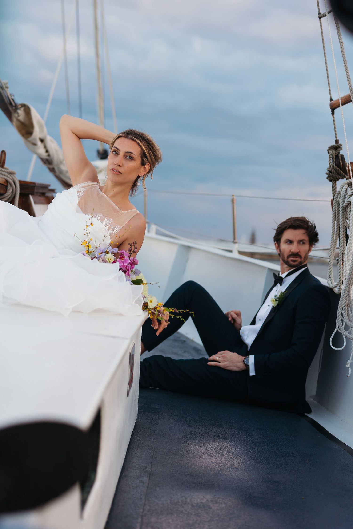 Bride and groom posing elegantly on a sailboat deck, bride holding a floral bouquet, groom in a tuxedo, cloudy sky backdrop.