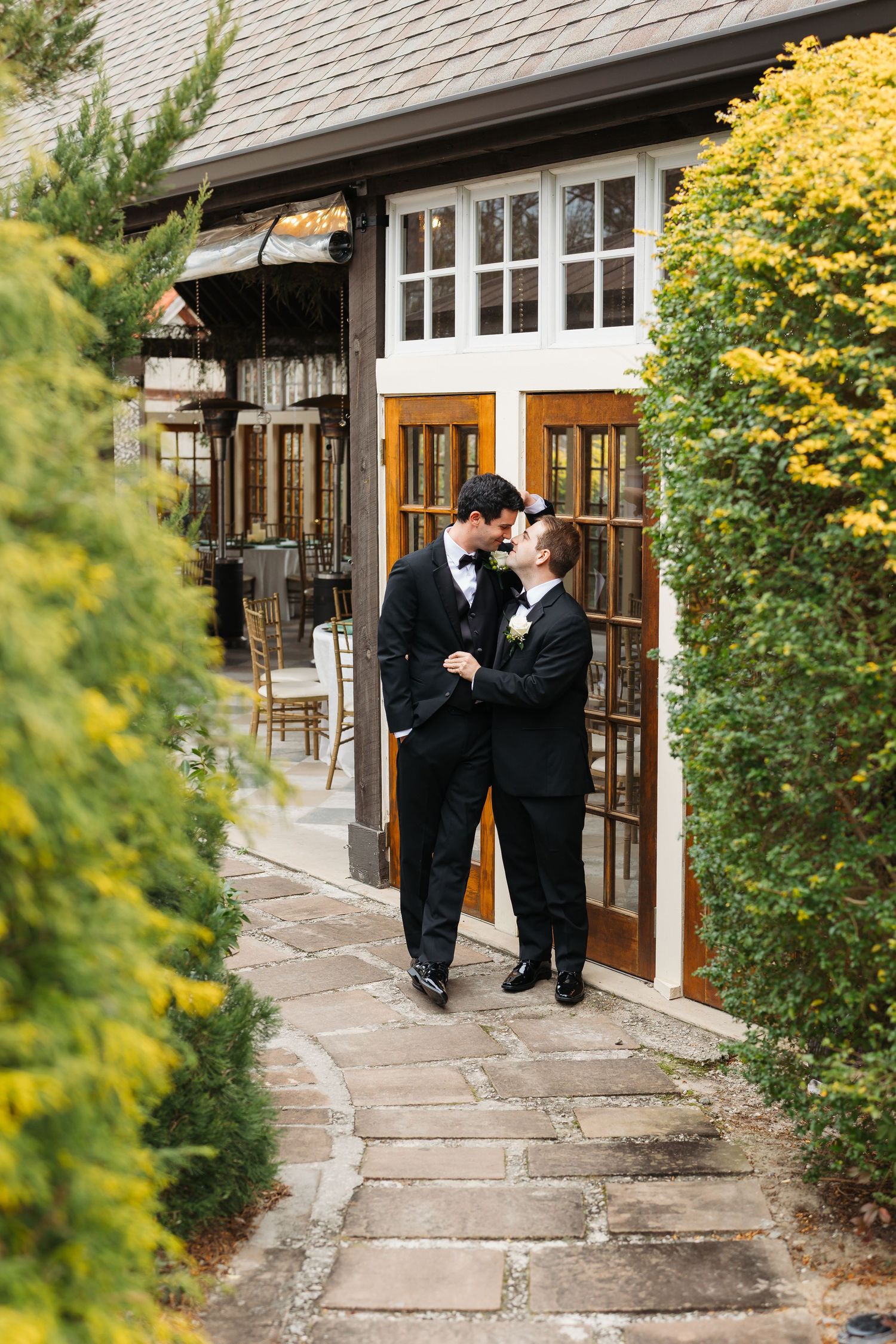 Two people in formal black attire share a tender moment on a stone path outside a rustic building with yellow foliage.