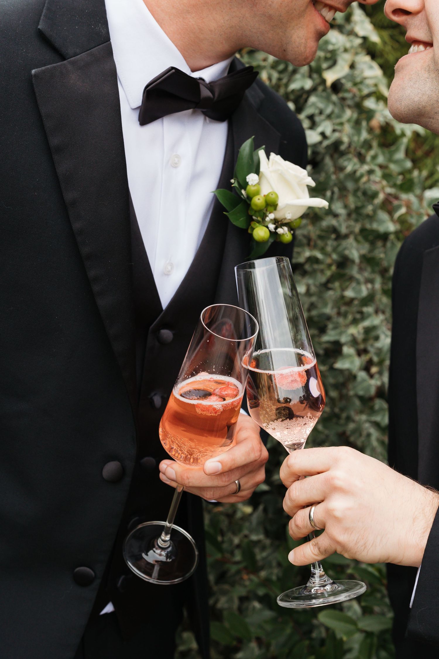Two people in formal attire toast with glasses of rose champagne, with one wearing a black tuxedo and white boutonniere.