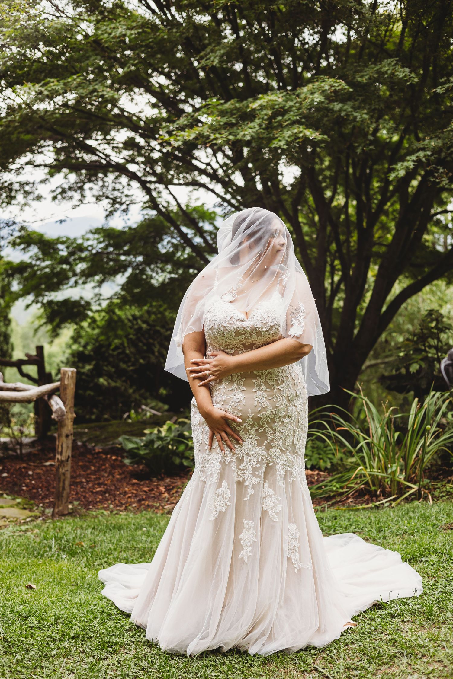 A flowing white lace wedding dress with short sleeves is displayed in a garden setting with lush green trees and grass.