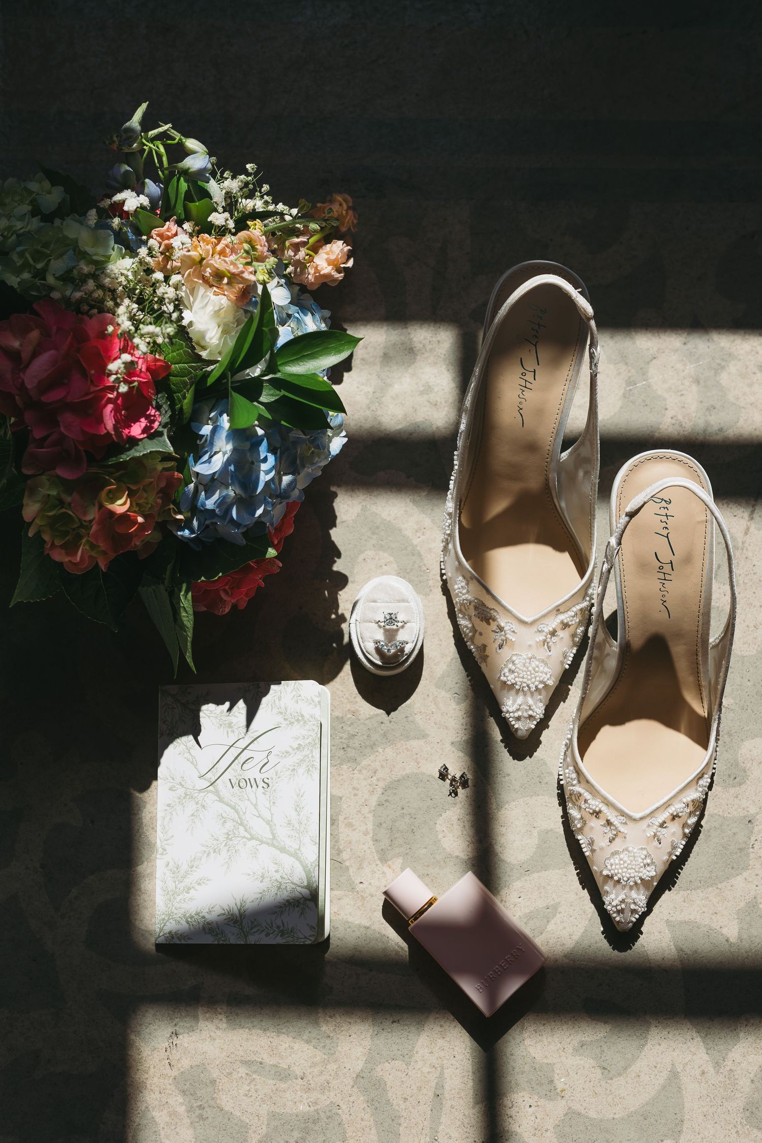 Elegant bridal flat lay featuring lace heels, floral bouquet, vow book, ring, and perfume on a dark textured surface.