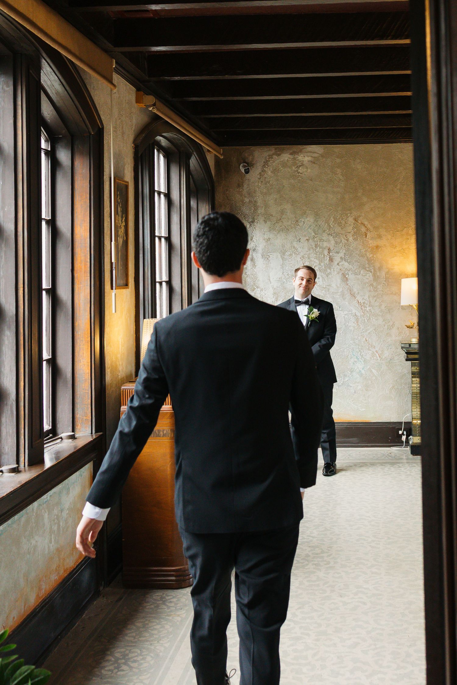 Two people in black tuxedos stand in an elegant hallway with large windows and exposed wooden beams.