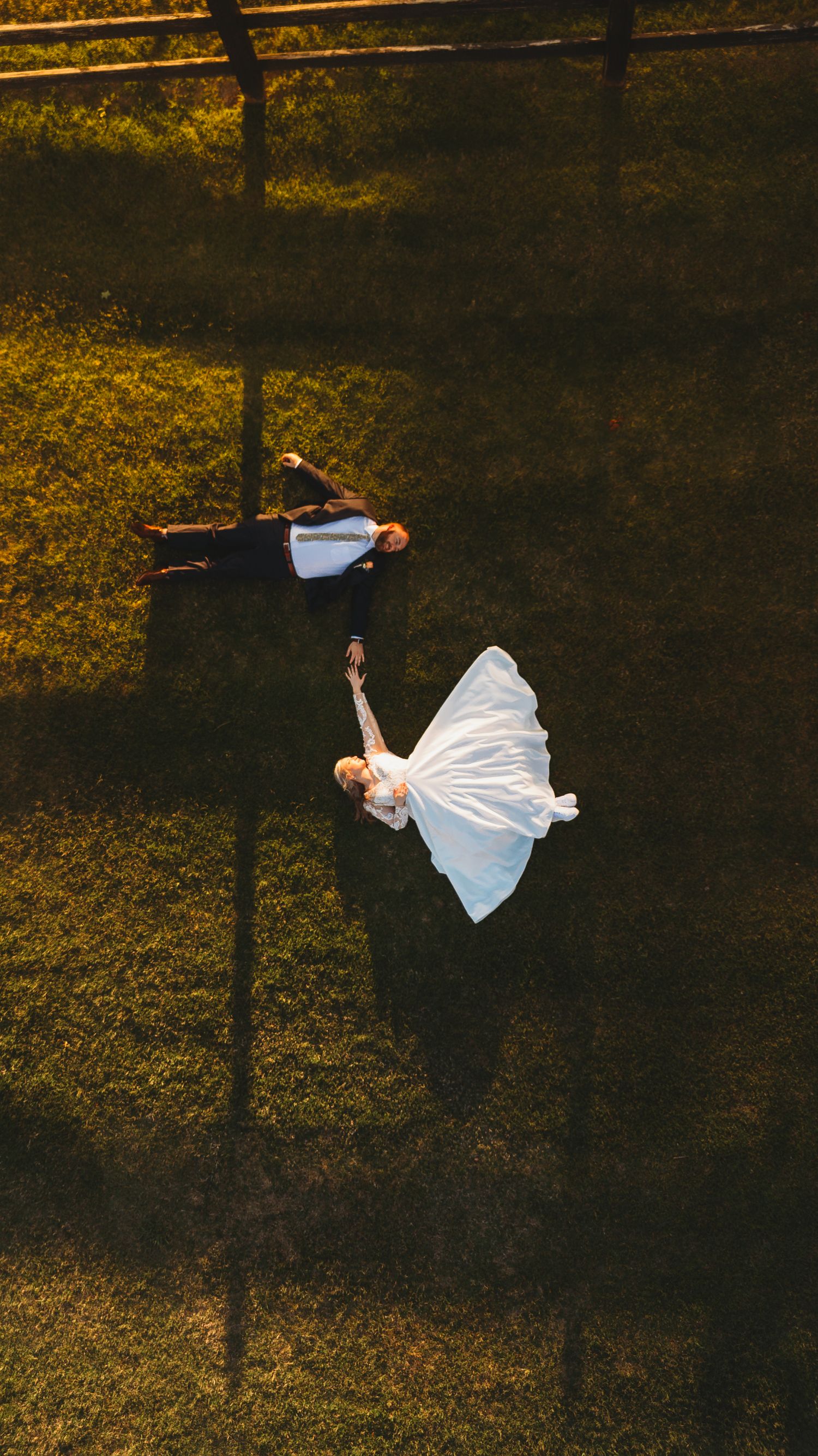 Aerial view of a couple lying on grass at sunset, with bride in white dress and groom in dark suit creating contrast.