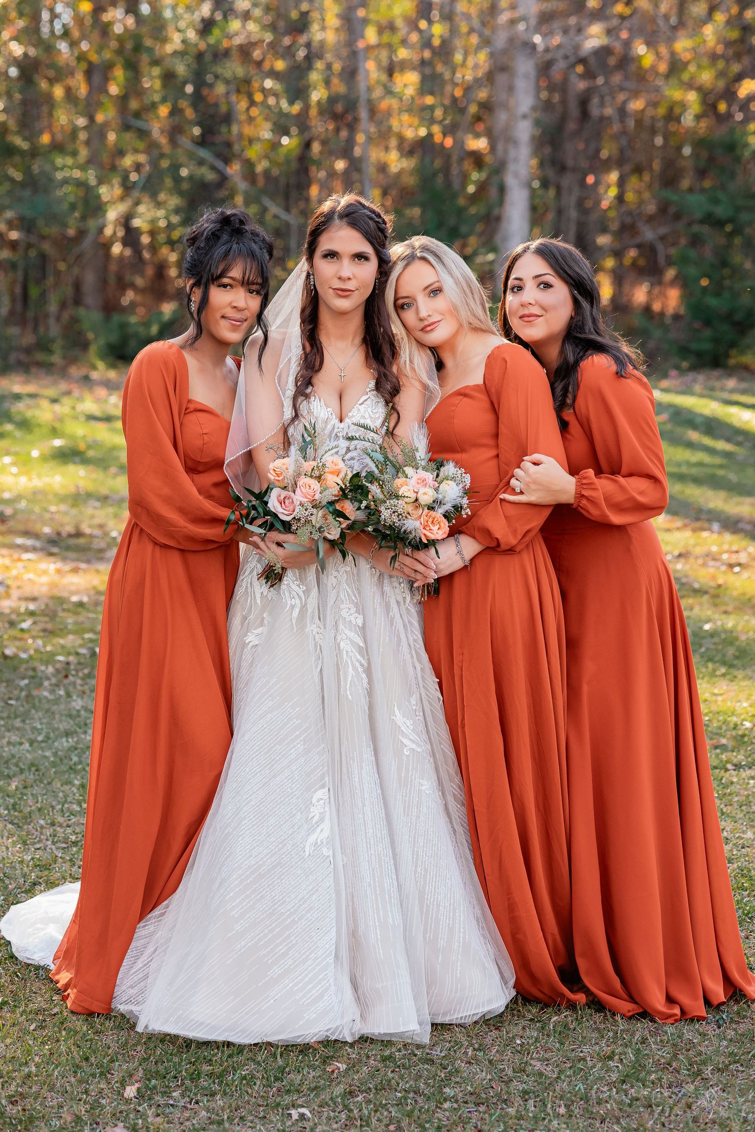A bride in a white lace gown stands with three bridesmaids wearing matching burnt orange dresses in an autumn setting.