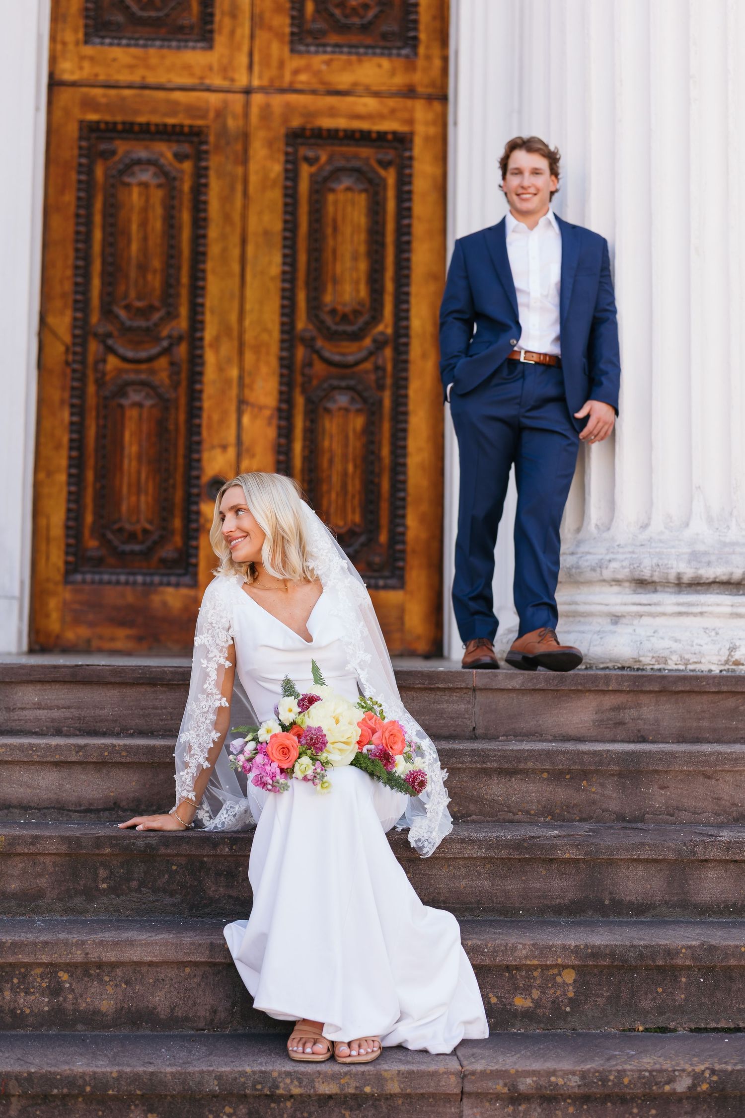 Bride in white gown holds colorful bouquet on steps while groom in navy suit leans against column behind her.