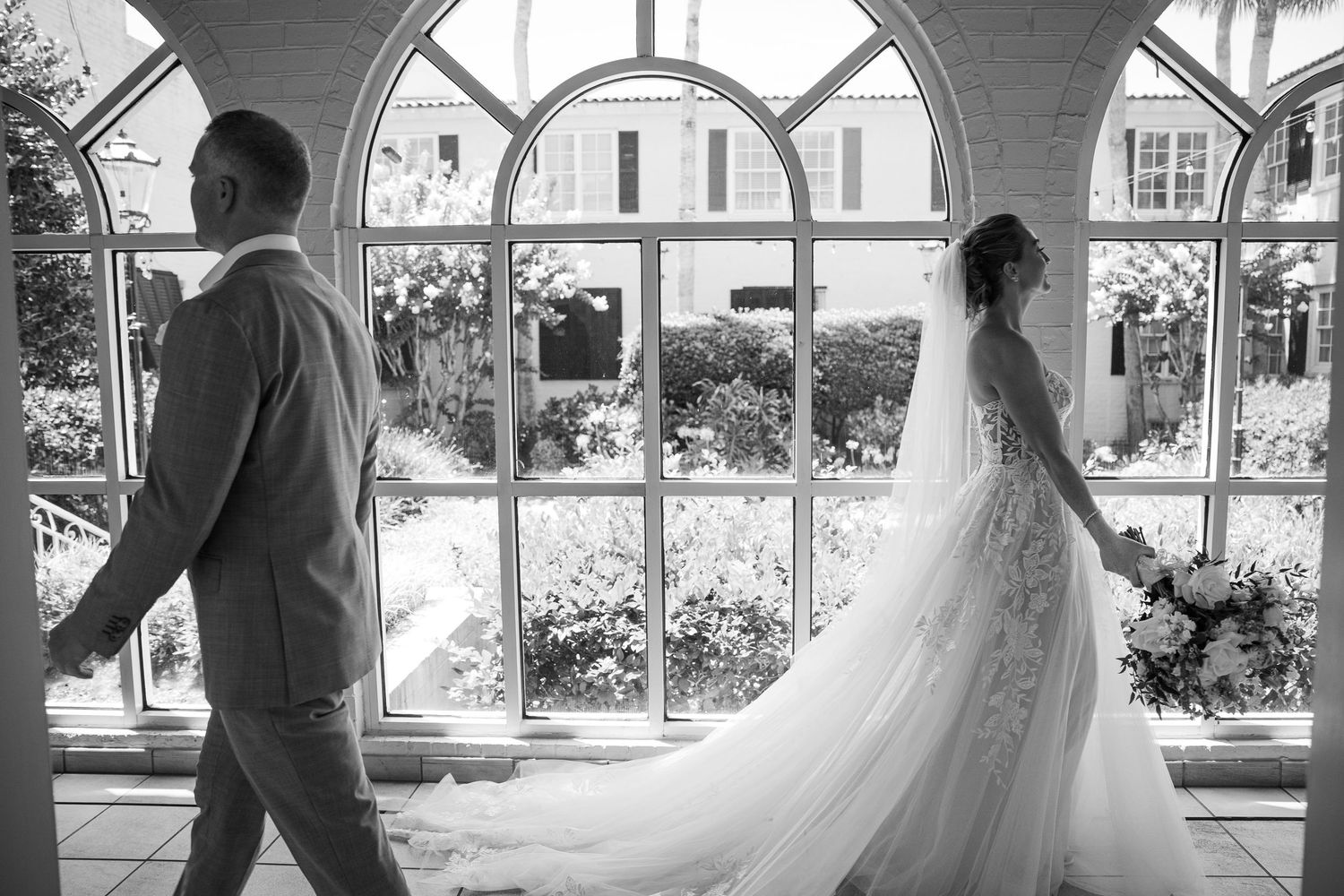 Black and white wedding photo showing bride in flowing gown and groom walking separately in elegant conservatory setting.